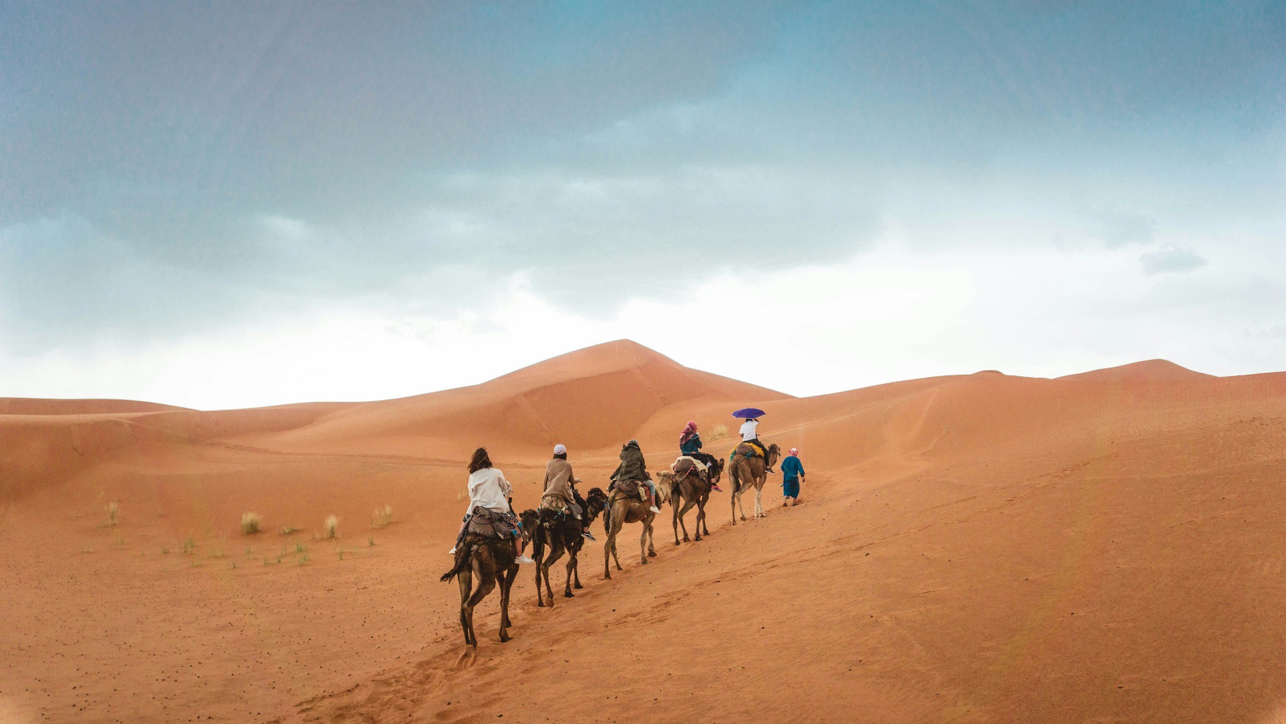 Group of people riding camels in desert with sand dunes and cloudy sky