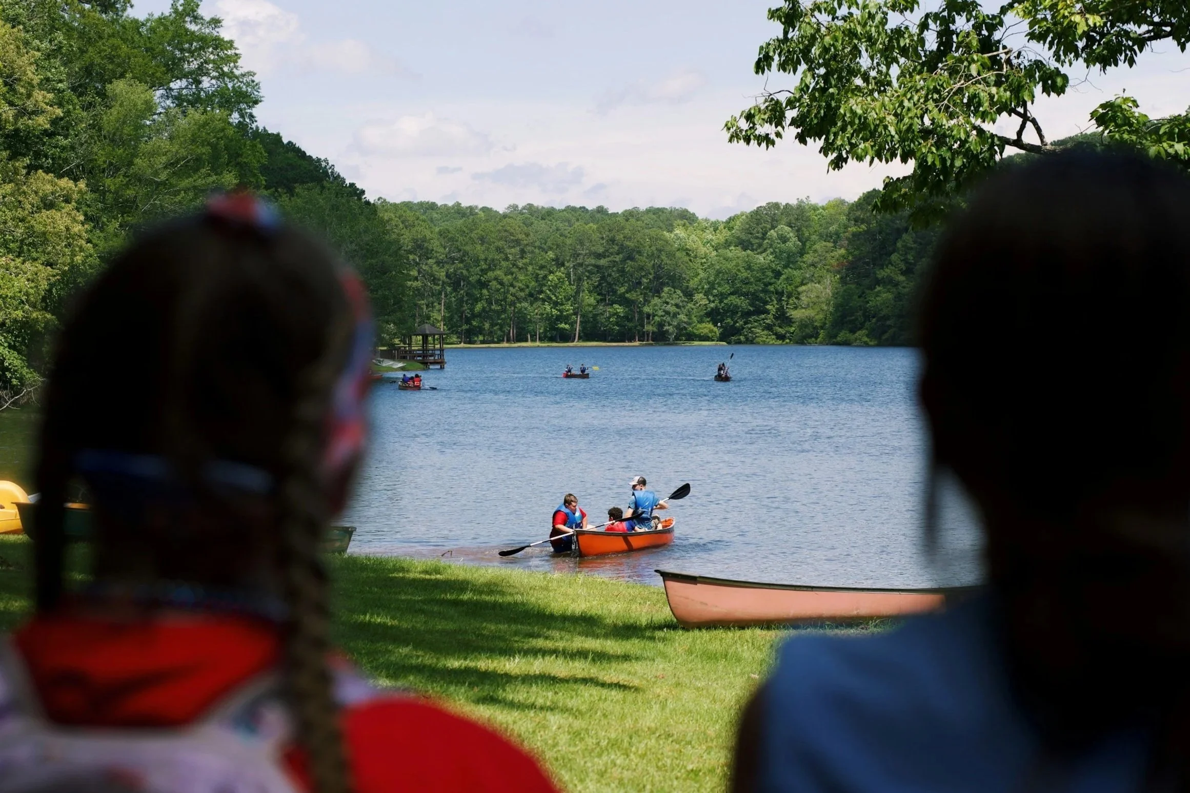 People canoeing on a lake surrounded by trees, viewed through two indistinct figures in the foreground.