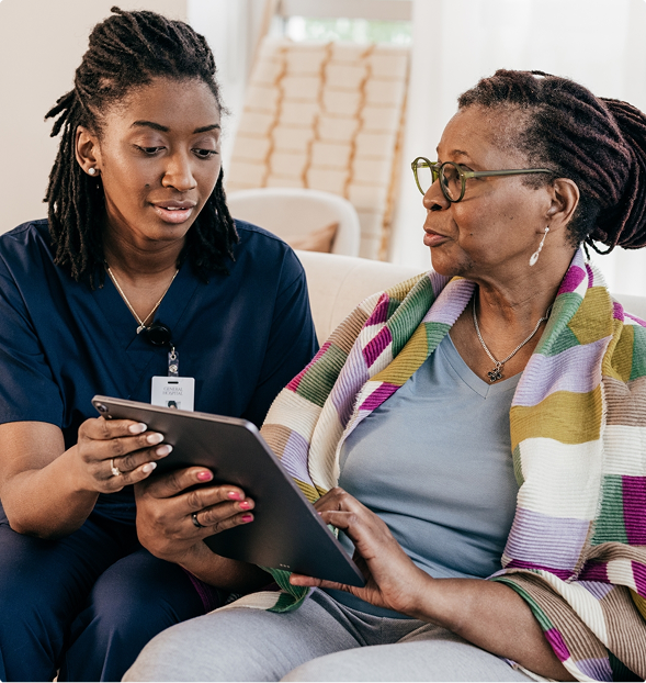 A young woman, possibly a healthcare worker, shows information on a tablet to an older woman in a cozy living room setting.