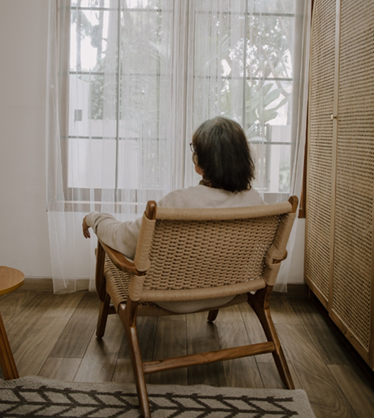 Person sitting on a wicker chair facing large window with sheer curtains, indoors with wooden flooring and furniture. In-home hospice care setting with comfortable wheelchair - Lotus Blossom North Georgia.