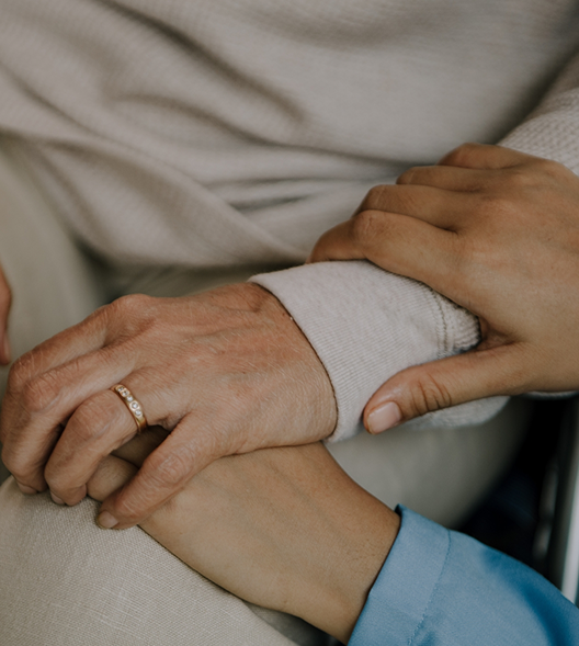 Close-up of two people holding hands, one male with a wedding ring, the other female with a plain blue shirt, conveying support and care.