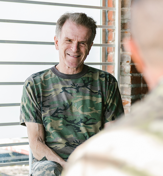 An elderly man smiling, sitting indoors near a window with horizontal blinds and a brick wall, wearing a camouflage T-shirt.