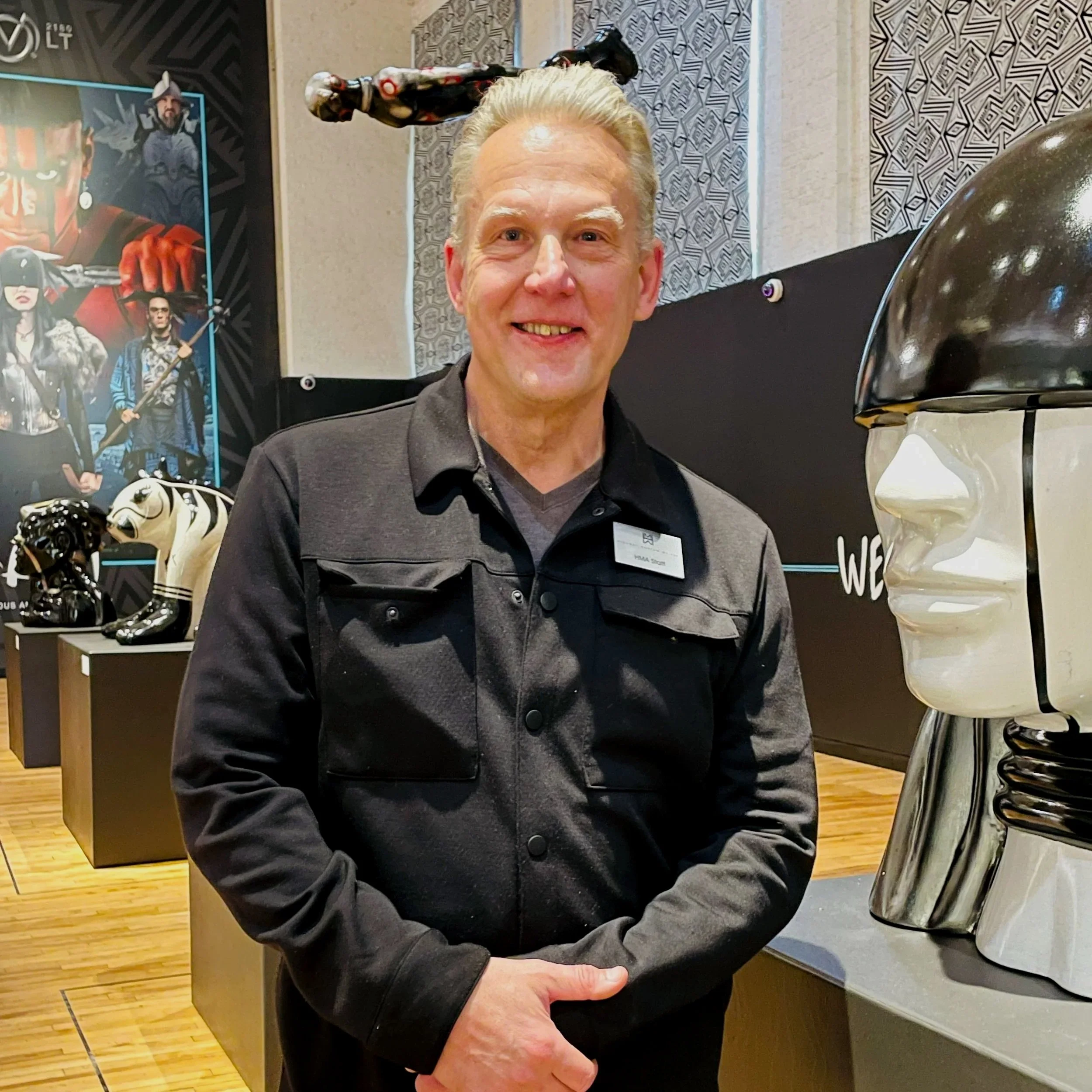 A man smiling at a museum exhibit featuring various art sculptures, including a large metallic head sculpture with a black band, and other animal-themed sculptures in the background.