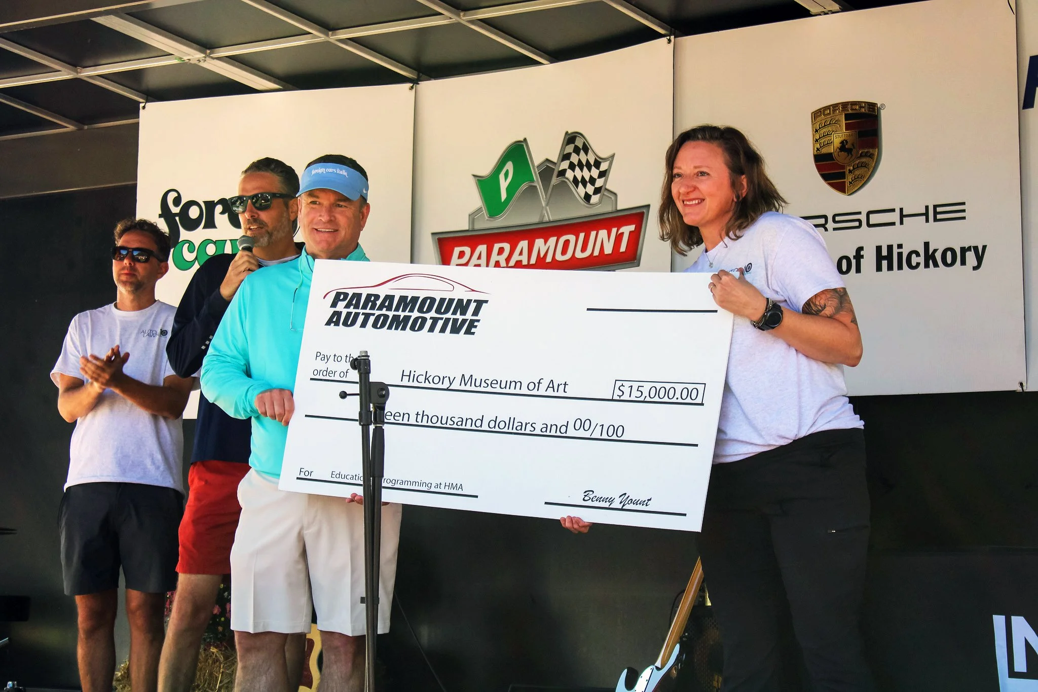 Group of people on stage during an award ceremony, one man holding a large check for $15,000 made out to Hickory Museum of Art, with logos of Porsche, Paramount, and other sponsors in the background.