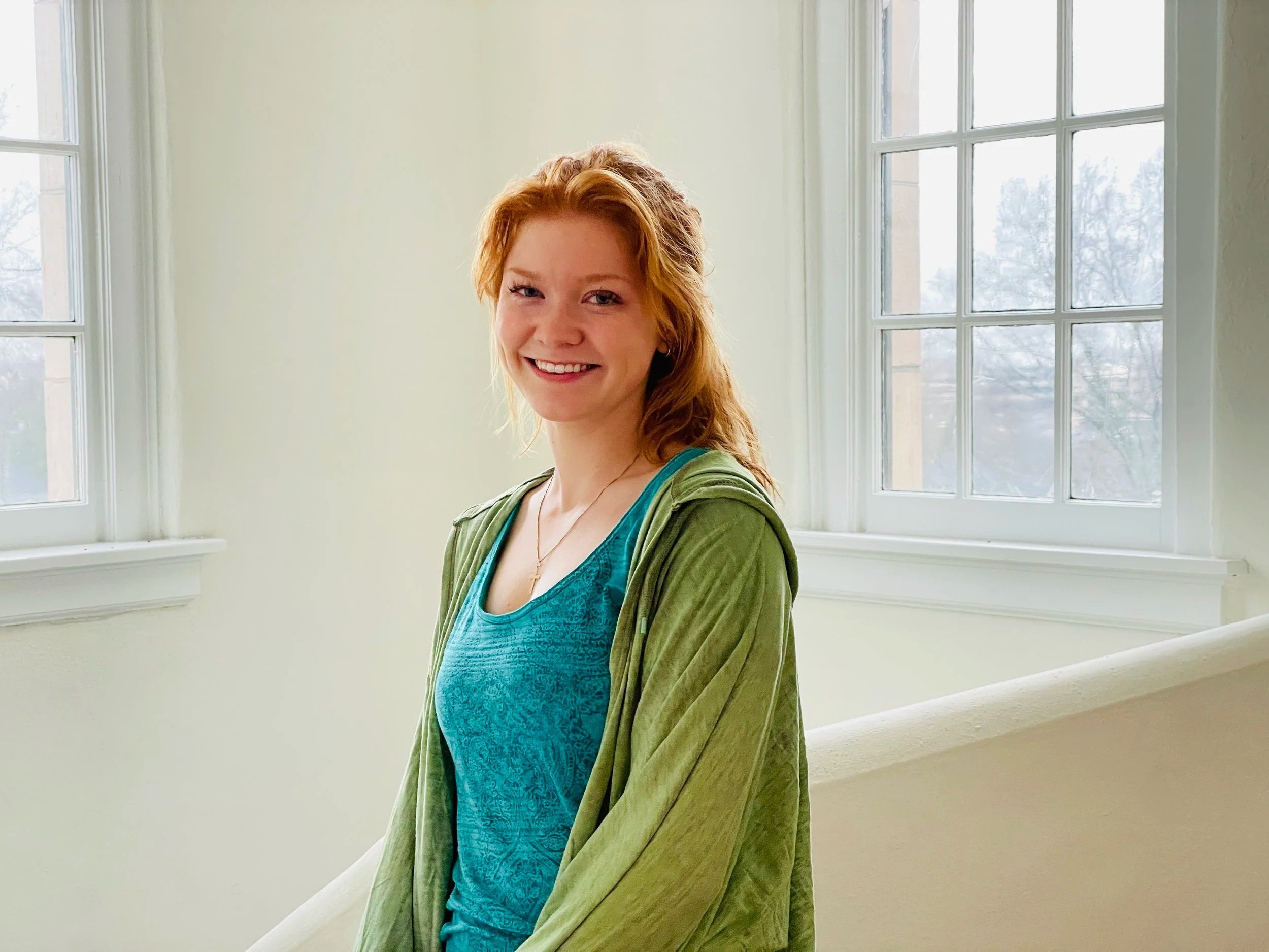 A young woman with red hair smiling in a brightly lit room with large windows.