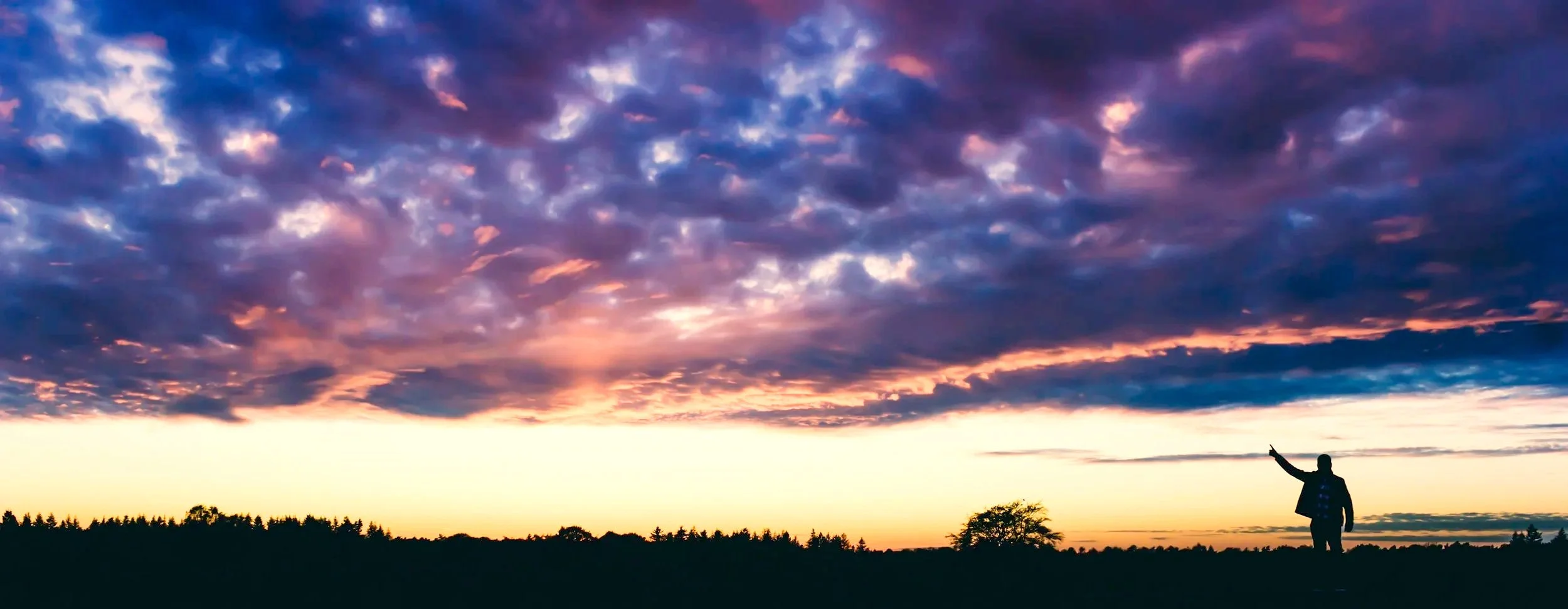 Silhouette of a person standing in a field pointing at a colorful sunset sky filled with clouds.