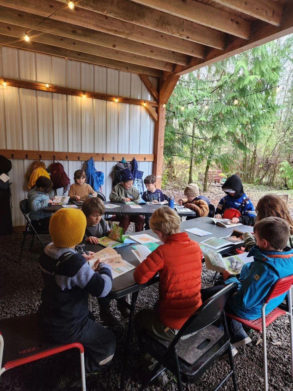 Two children, a girl and a boy, are in a tree. The girl is sitting on a branch, writing in a notebook, while the boy is lying on a branch, looking at a notepad. There are backpacks hanging on the tree and on the ground.