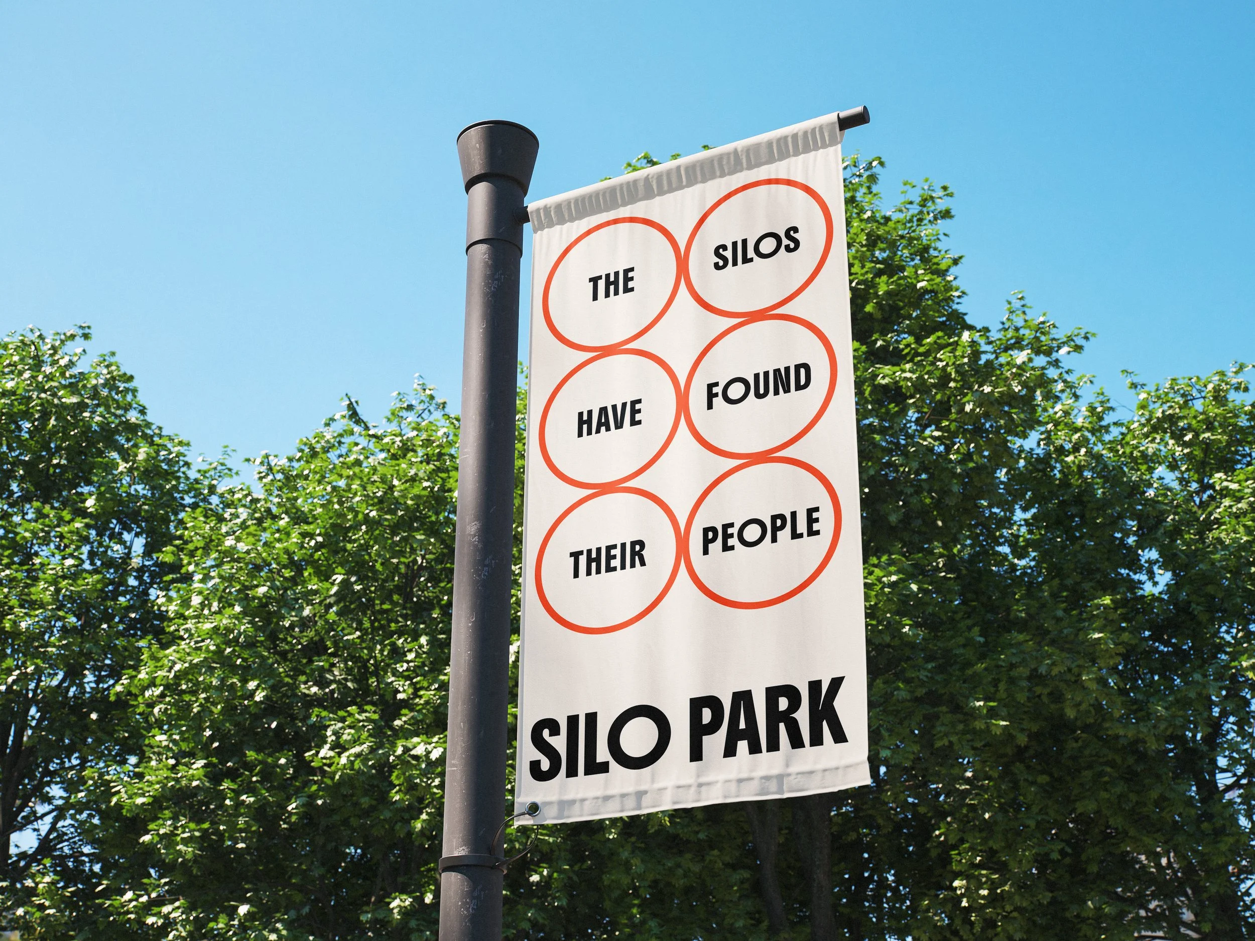 A sign with a white background and orange circles, written with black letters, reads 'THE SILOS HAVE FOUND THEIR PIPE SILO PARK'. The sign is attached to a metal pole, with green trees and a clear blue sky in the background.