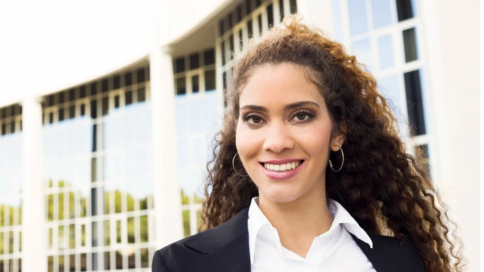 A young woman with curly hair, wearing a black blazer and white shirt, smiling outdoors in front of a modern building with glass windows.