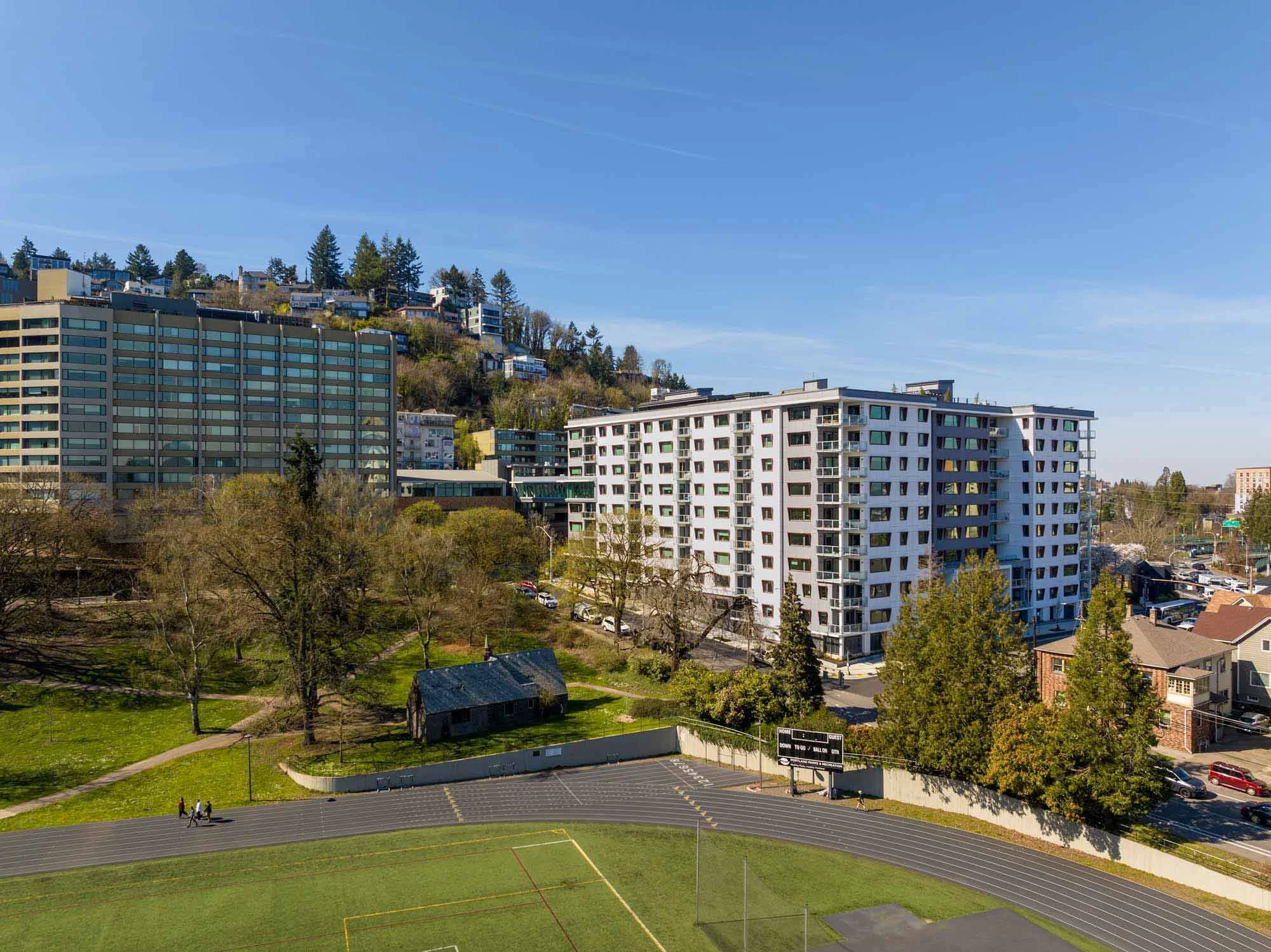 Cityscape with high-rise apartment buildings on a hillside, trees, a small park, and a running track in the foreground under a clear blue sky.