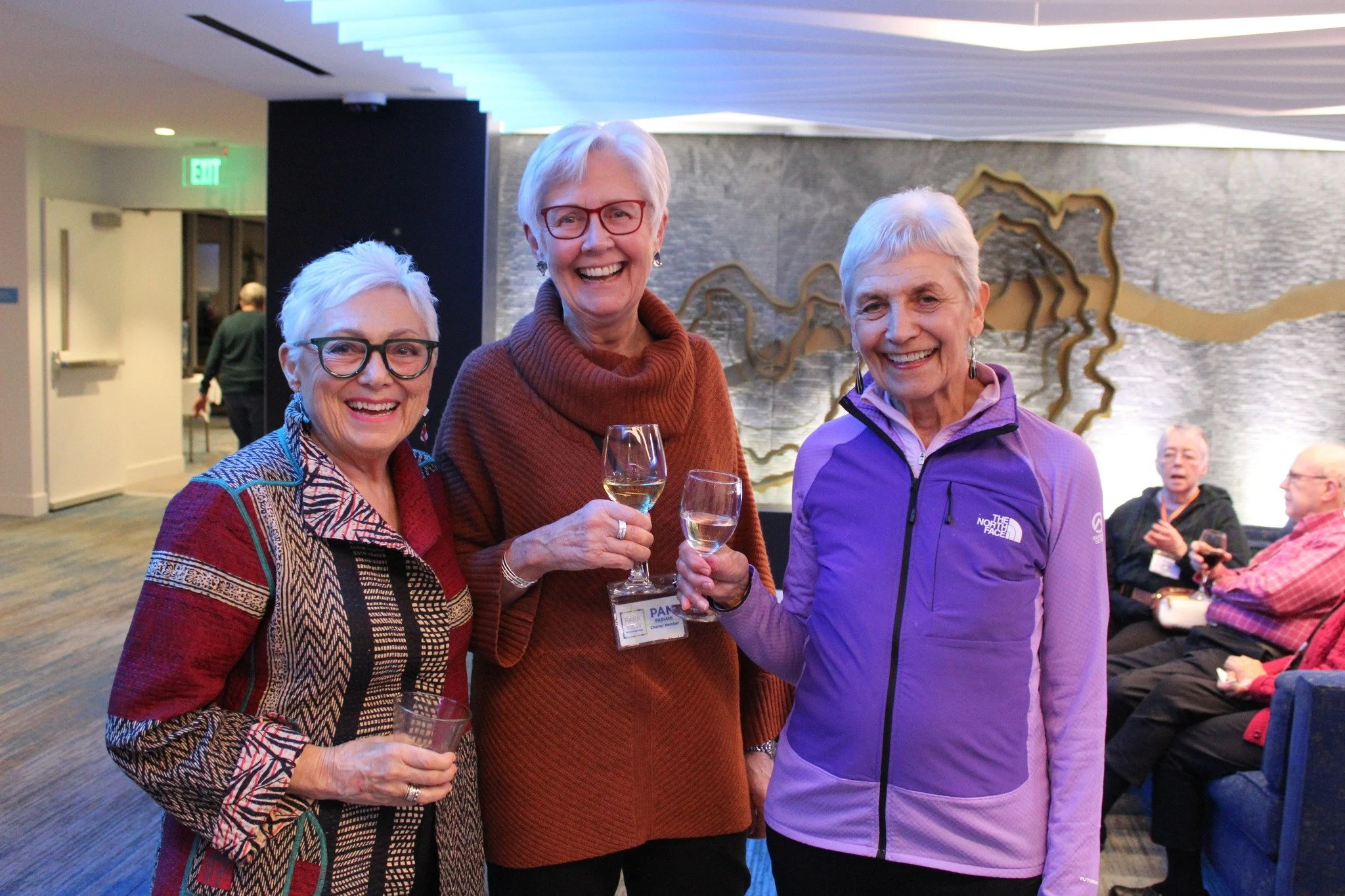 Three women smiling and holding glasses of white wine at an indoor event, with other guests seated in the background.