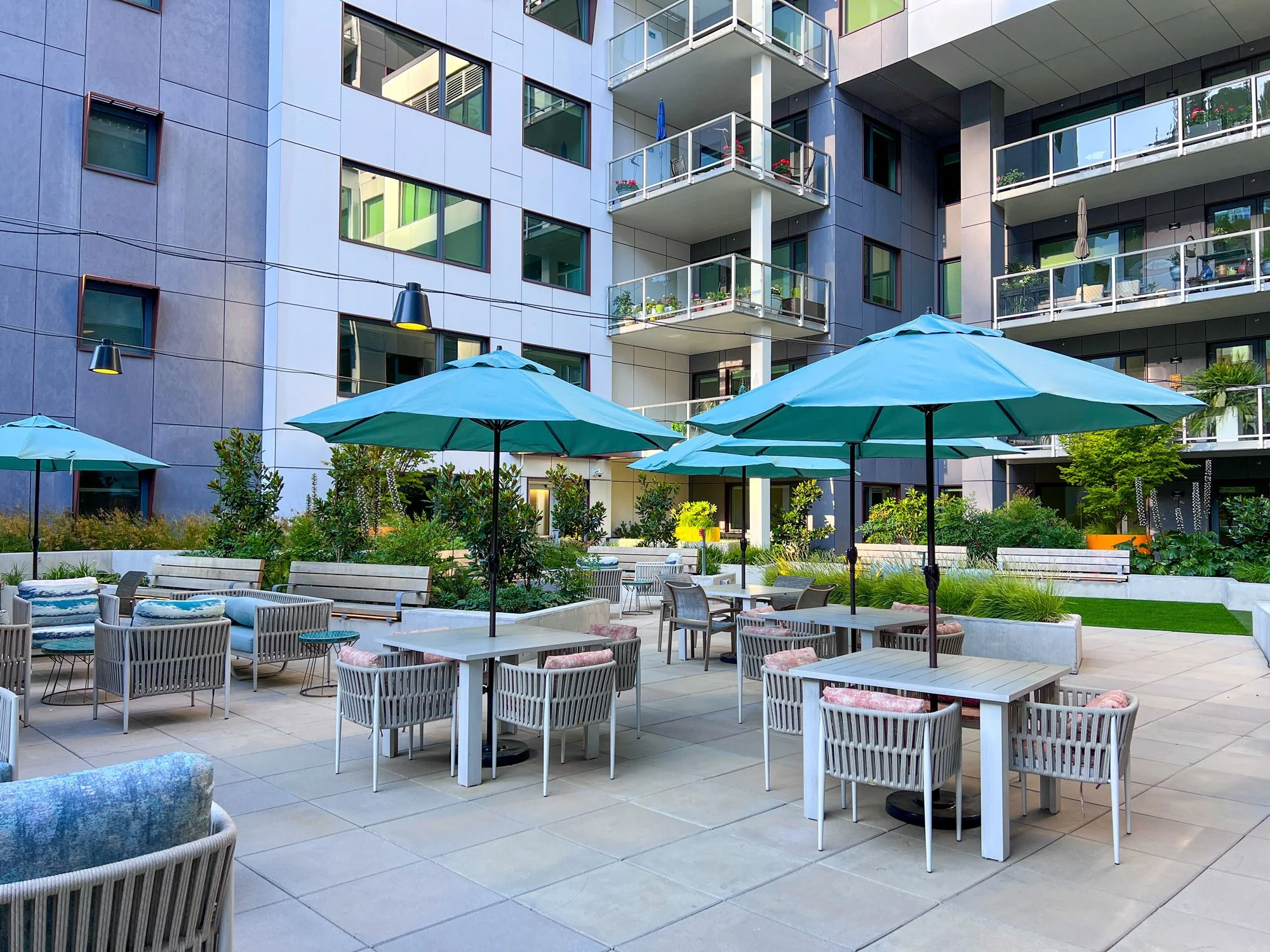 An outdoor patio area with tables, chairs, and large blue umbrellas, surrounded by greenery and modern apartment buildings.