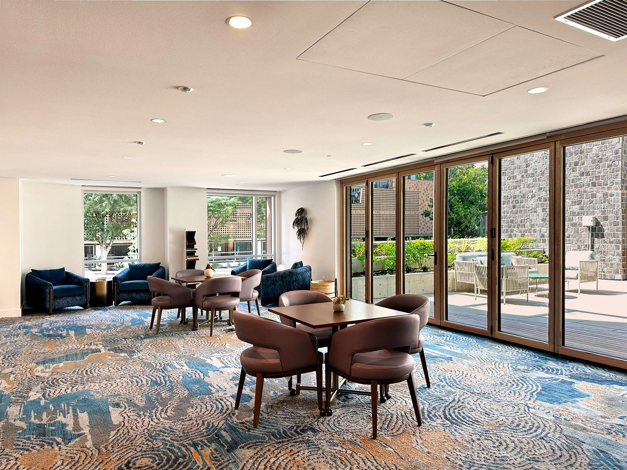 Modern, well-lit lounge with large windows, seating areas with blue and taupe chairs, a decorative rug, and an outdoor patio visible through glass sliding doors.