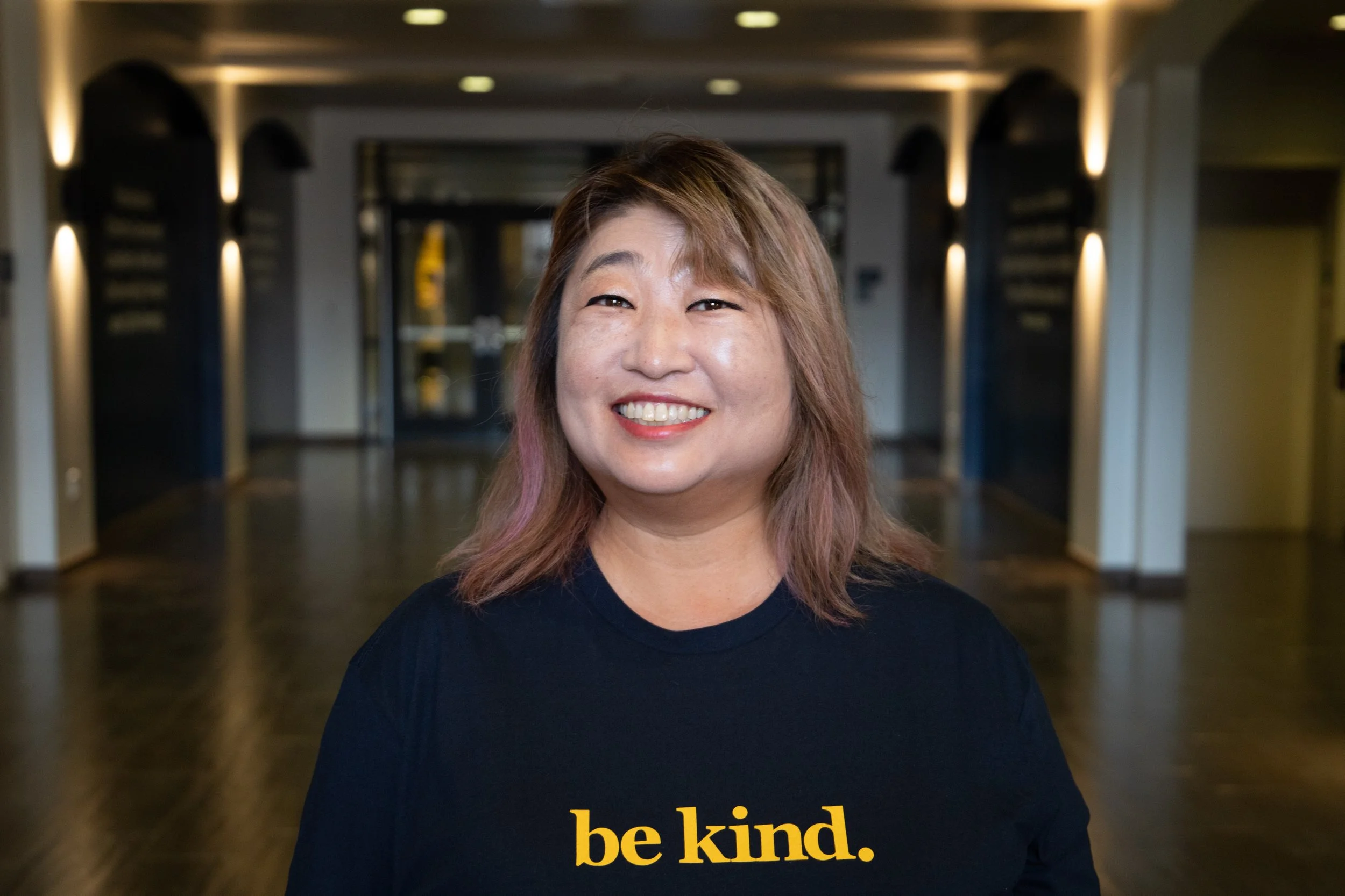 A woman with shoulder-length brown and pink hair smiling, wearing a navy blue shirt with yellow text that reads 'be kind.' in a modern indoor setting with warm lighting.