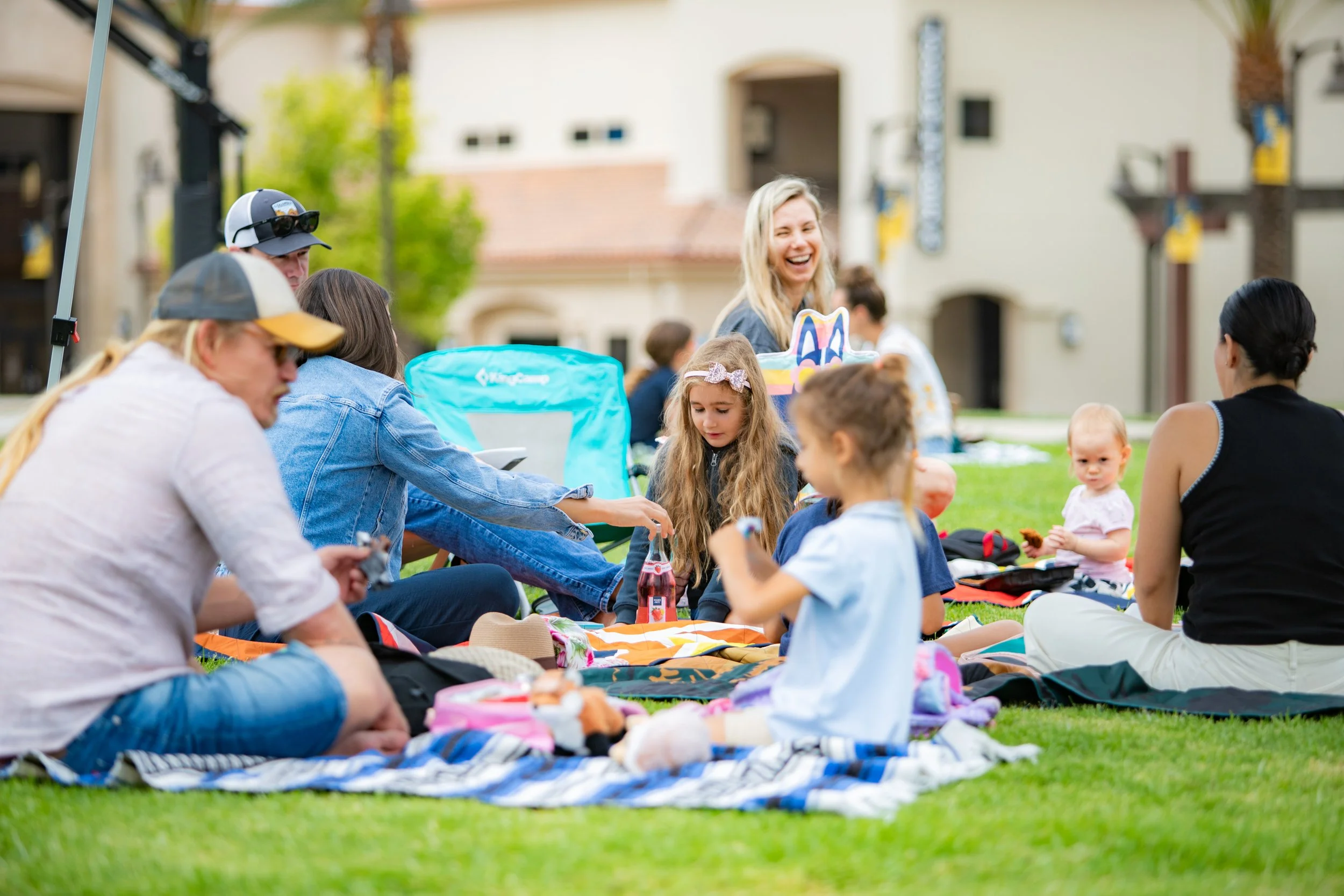 A group of people, including children and adults, gathered on a grassy lawn outdoors for a daytime event. They are sitting on blankets and engaging in activities, with some smiling and chatting. There are trees and residential buildings in the background, and the atmosphere appears cheerful and festive.