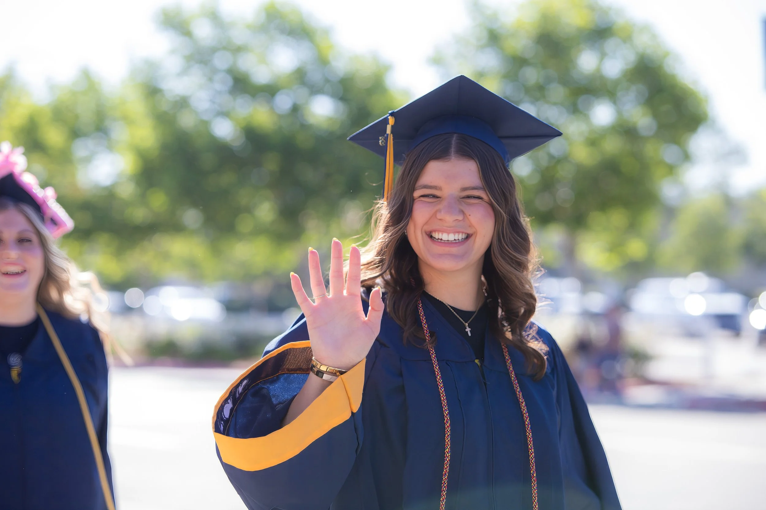 A young woman in a diploma gown and cap waving at the camera during a graduation ceremony outdoors on a sunny day, with trees in the background.