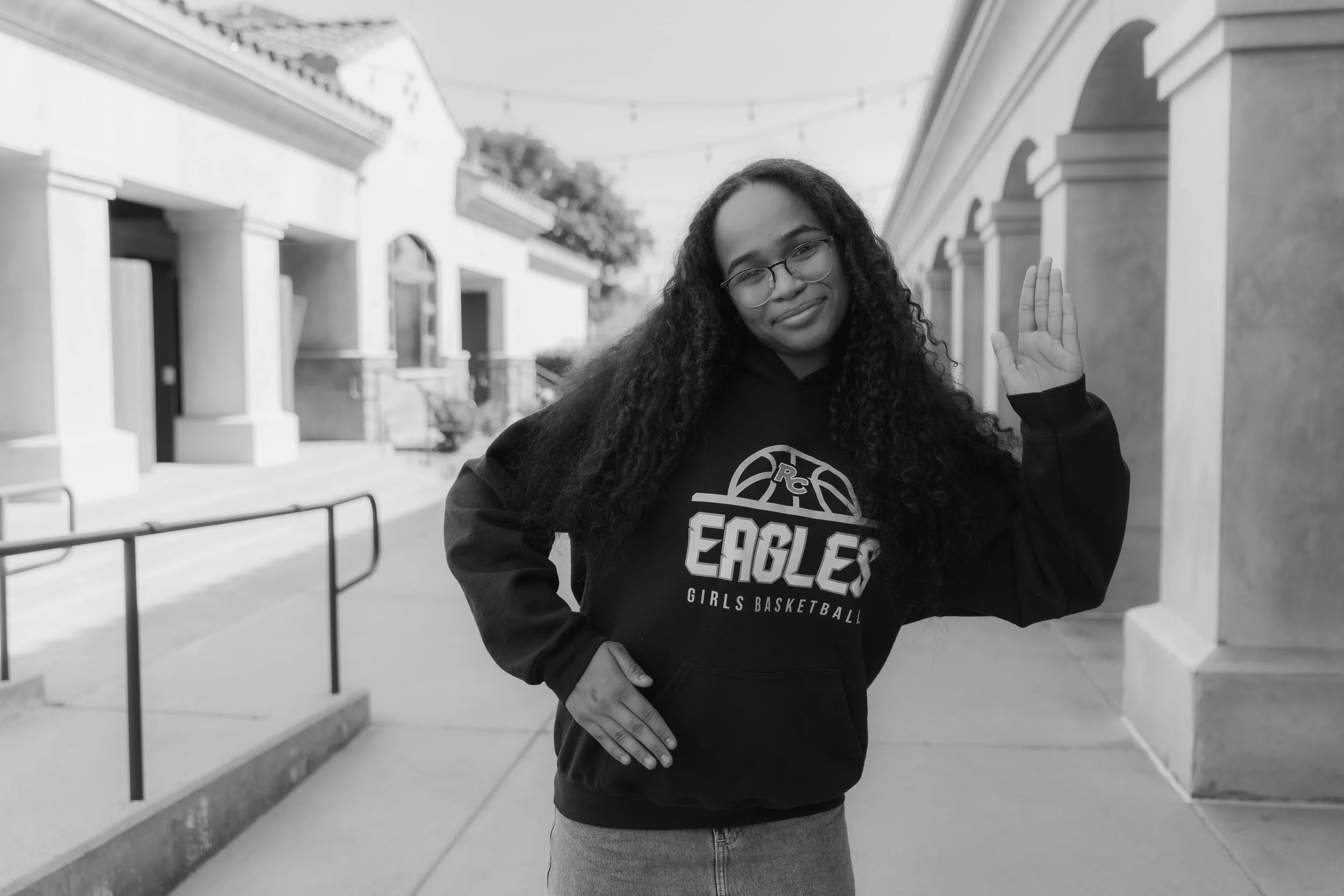 Young woman with glasses, long brown hair, smiling, wearing a navy blue 'Eagles' t-shirt, standing against a beige wall.