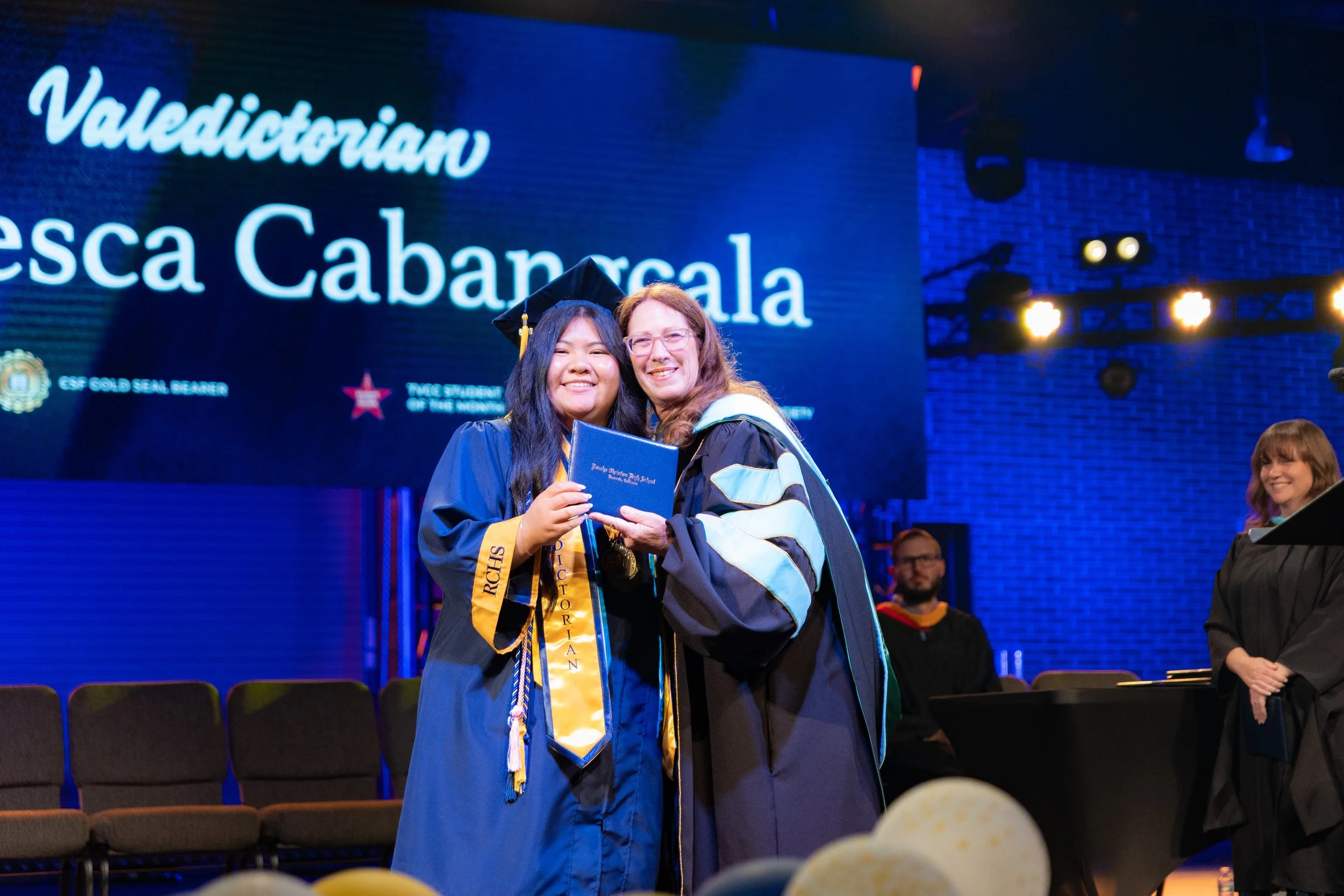 A graduation ceremony at the University of California showing three people smiling, including a woman in a graduation cap and gown holding a diploma, with a large university banner in the background.