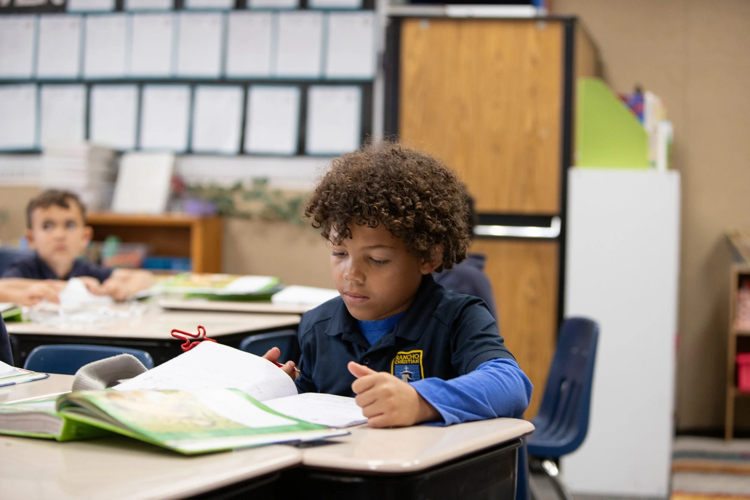 A young girl with a pink bow in her hair reading a yellow book in a classroom.