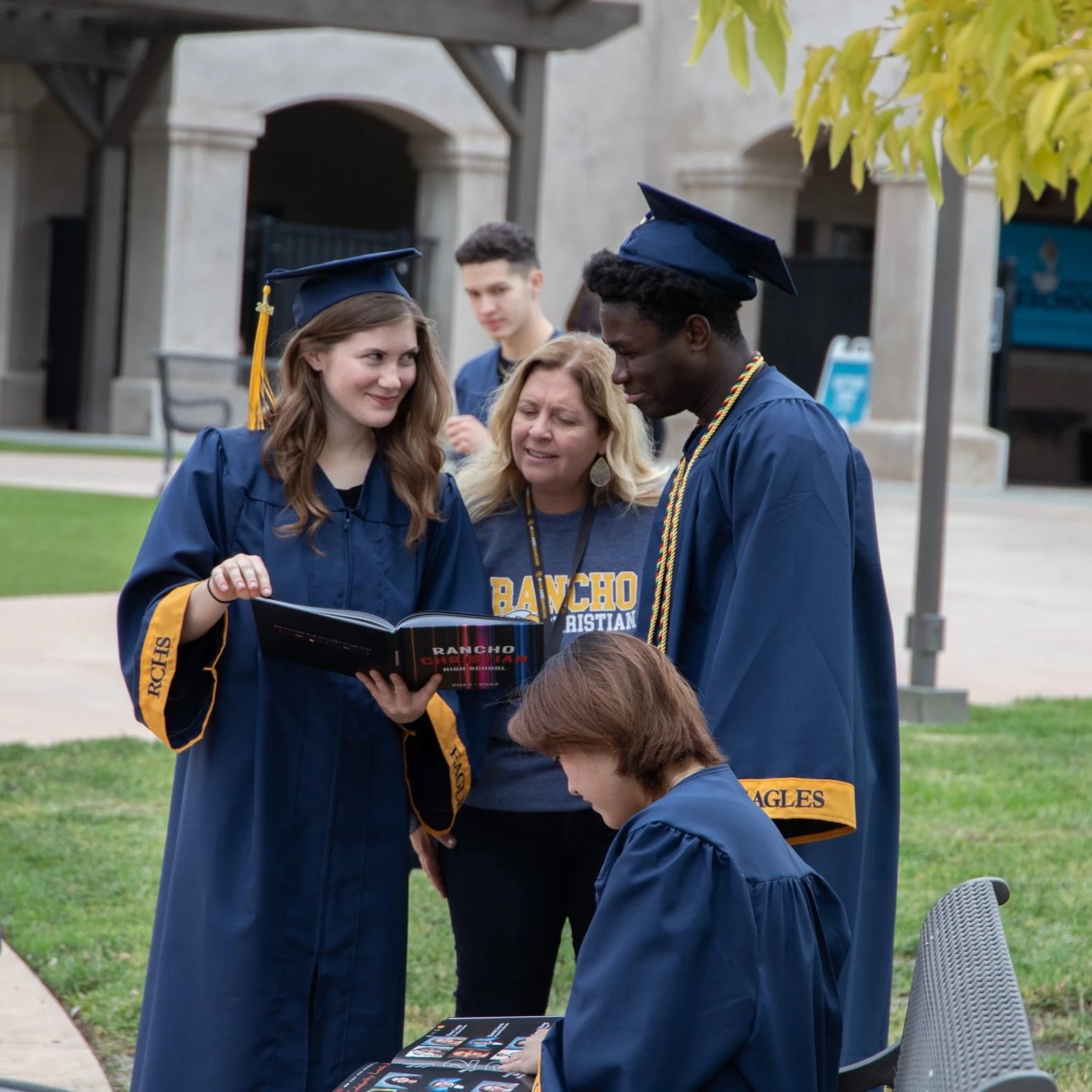 Three students standing in a school hallway, smiling. The female student on the left is wearing a black t-shirt with yellow text and black pants. The male student in the middle is wearing a black jacket and blue jeans. The male student on the right is wearing a gray hoodie and black pants, carrying a backpack. They are standing close with their arms around each other.