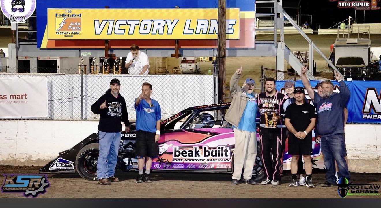 Group of six people celebrating in front of a pink racing car at a dirt track raceway, with a large sign reading 'Victory Lane' and various sponsor banners in the background.
