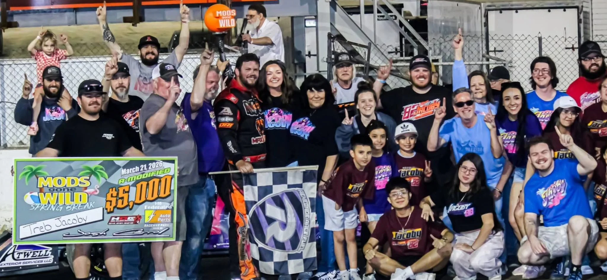 A large group of race car drivers, crew members, and children celebrating on a race track after winning a race. They are smiling, holding up one finger, and standing behind a checkered flag with a large check for $5,000. The scene includes a banner with race details and a crowd in the background.