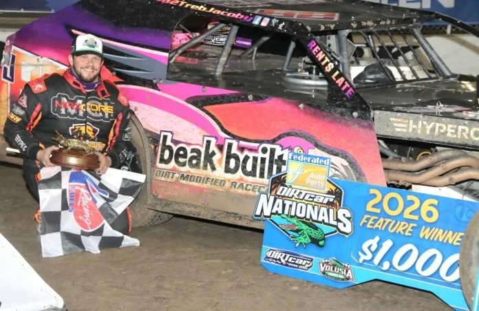 A race car driver in a black and red racing suit holding a trophy and checkered flag, standing beside a dirt track race car with sponsor decals, next to a large blue and yellow prize check for $1,000.