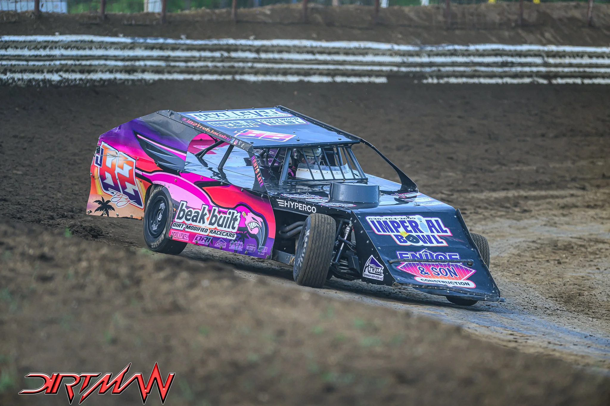 A colorful dirt track race car on a dirt track with advertisements and sponsor logos on its body, including 'beat built,' 'Imperial Bowl,' and 'J & Son Construction.' The car is in motion on a dirt race track with a blurred background.