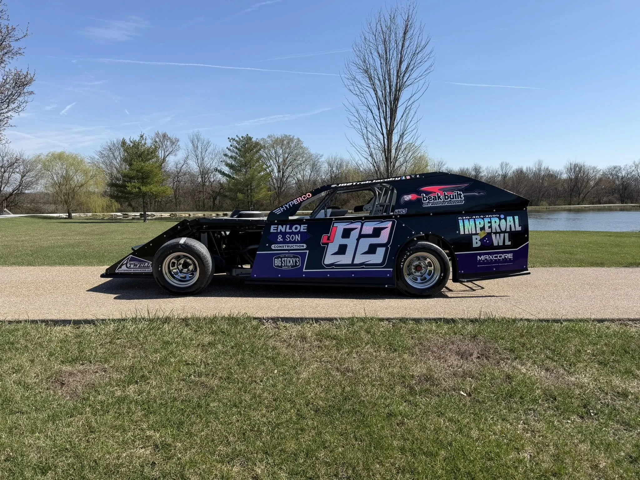 A race car with the number 26 on its side parked on a grassy area near a body of water with trees in the background and a blue sky overhead.
