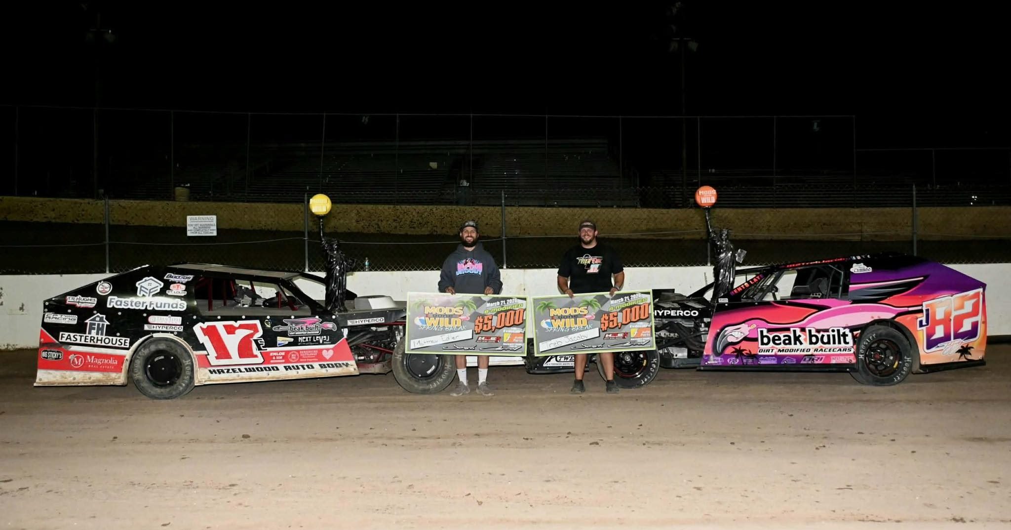 Two race cars on a dirt track with two men standing between them holding large checks for $5,000, and trophies on their cars, at night.