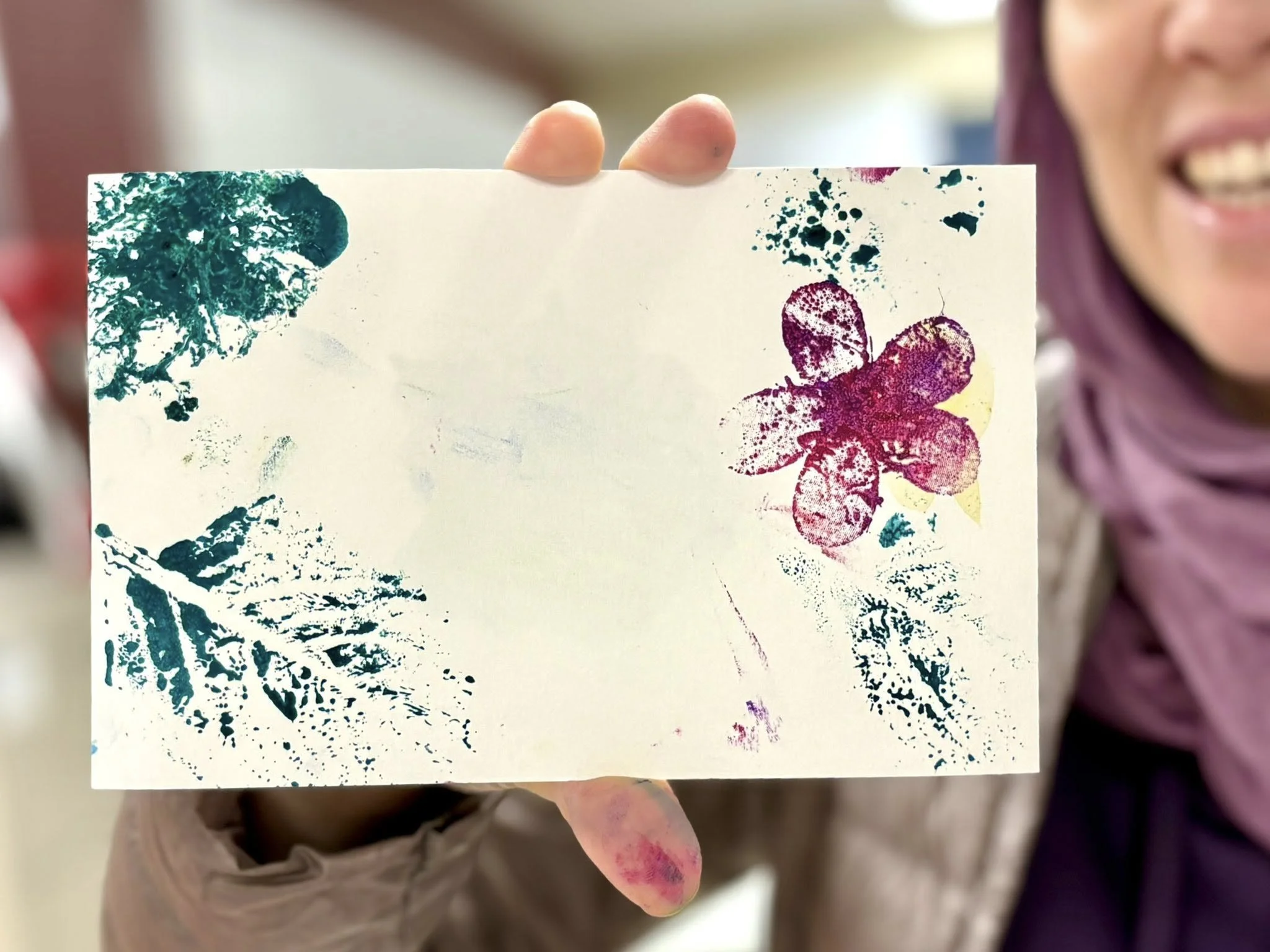 Une femme tient une carte blanche avec des impressions colorées de fleurs, de feuilles et de taches de peinture en vert, rose, violet et jaune.
