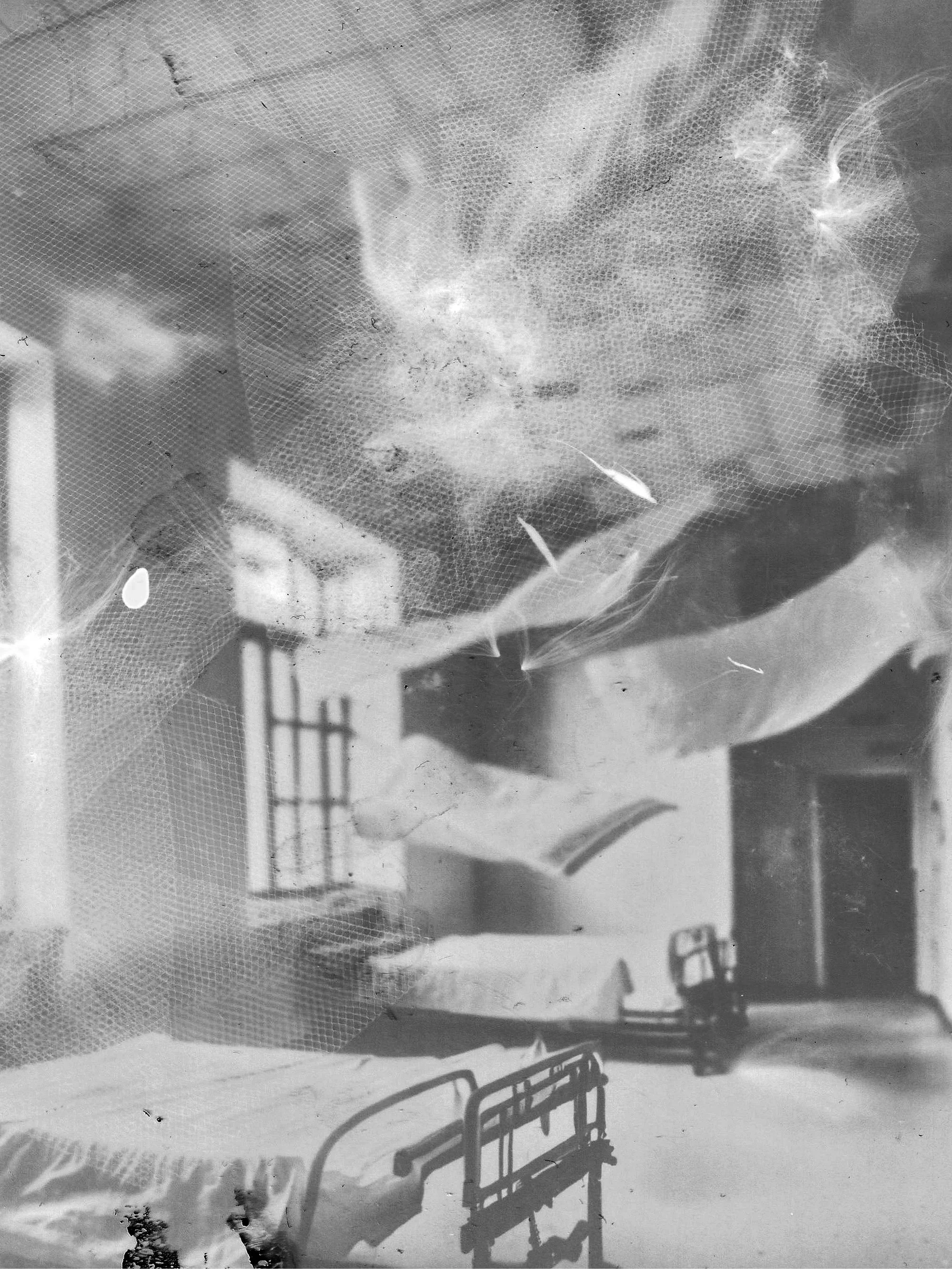 Une photo en noir et blanc d'une chambre avec deux lits séparés par une table de chevet, des fenêtres ouvertes laissant entrer de la lumière, et un mur blanc