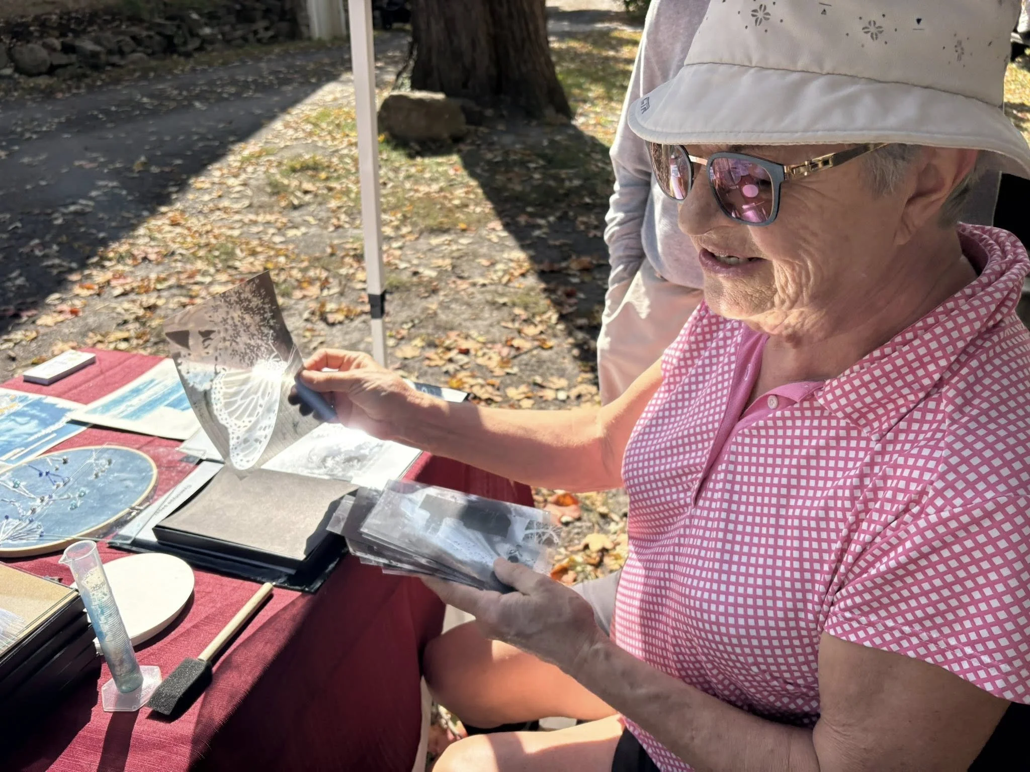 Une femme âgée portant une chemise rose à carreaux, des lunettes de soleil et une casquette blanche, regarde des photos ou des cartes dans ses mains, assise à une table avec divers objets à l'extérieur, parmi des feuilles mortes et un arbre.