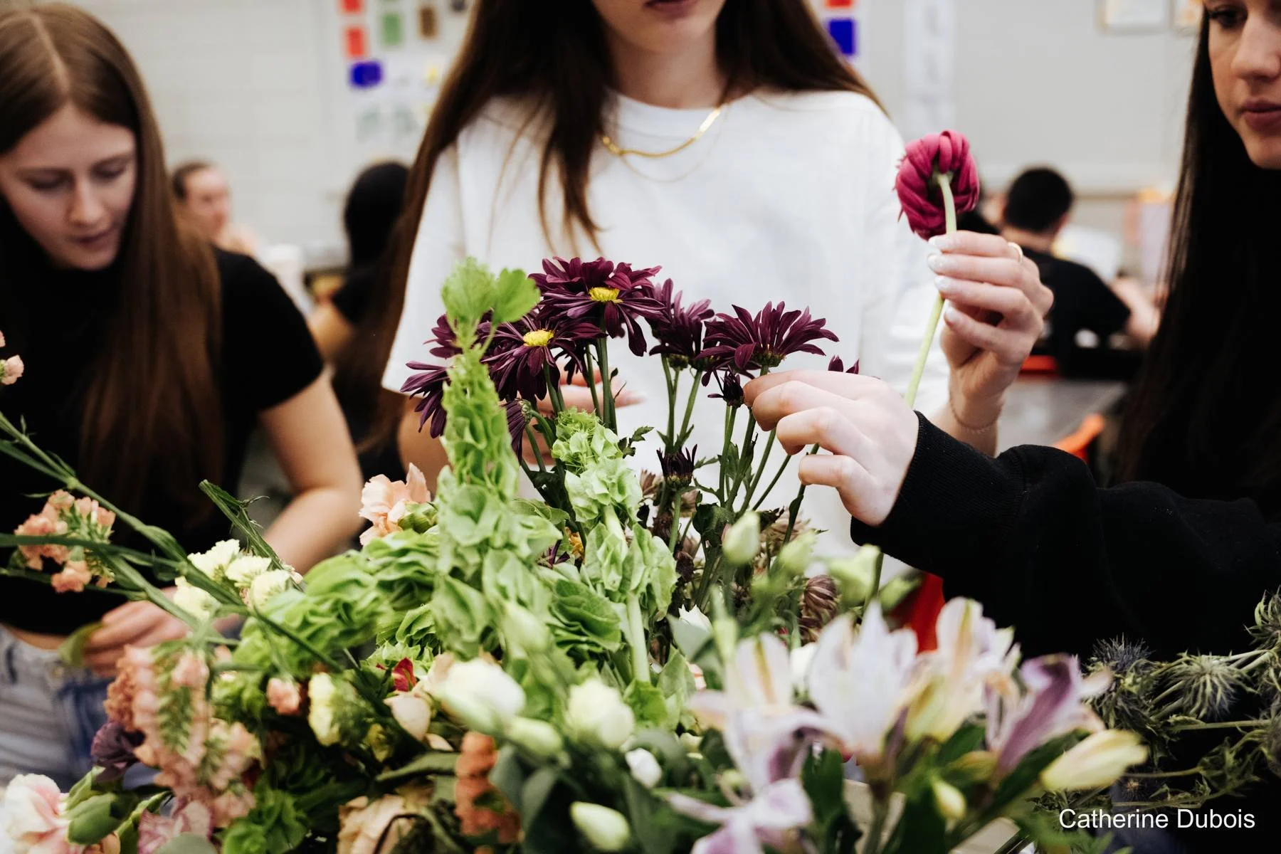 Groupe de personnes réunies autour d'un arrangement floral avec des fleurs violettes, roses et blanches, dans un environnement intérieur, probablement un atelier de composition florale.
