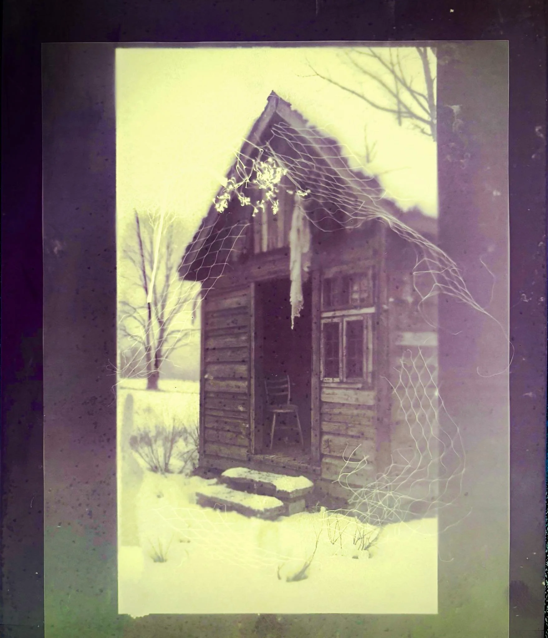 Une vieille cabane en bois dans un paysage enneigé, avec un fauteuil à l'intérieur visible par la porte ouverte, entourée d'arbres sans feuilles et d'un filet de pêche abandonné sur la cabane.