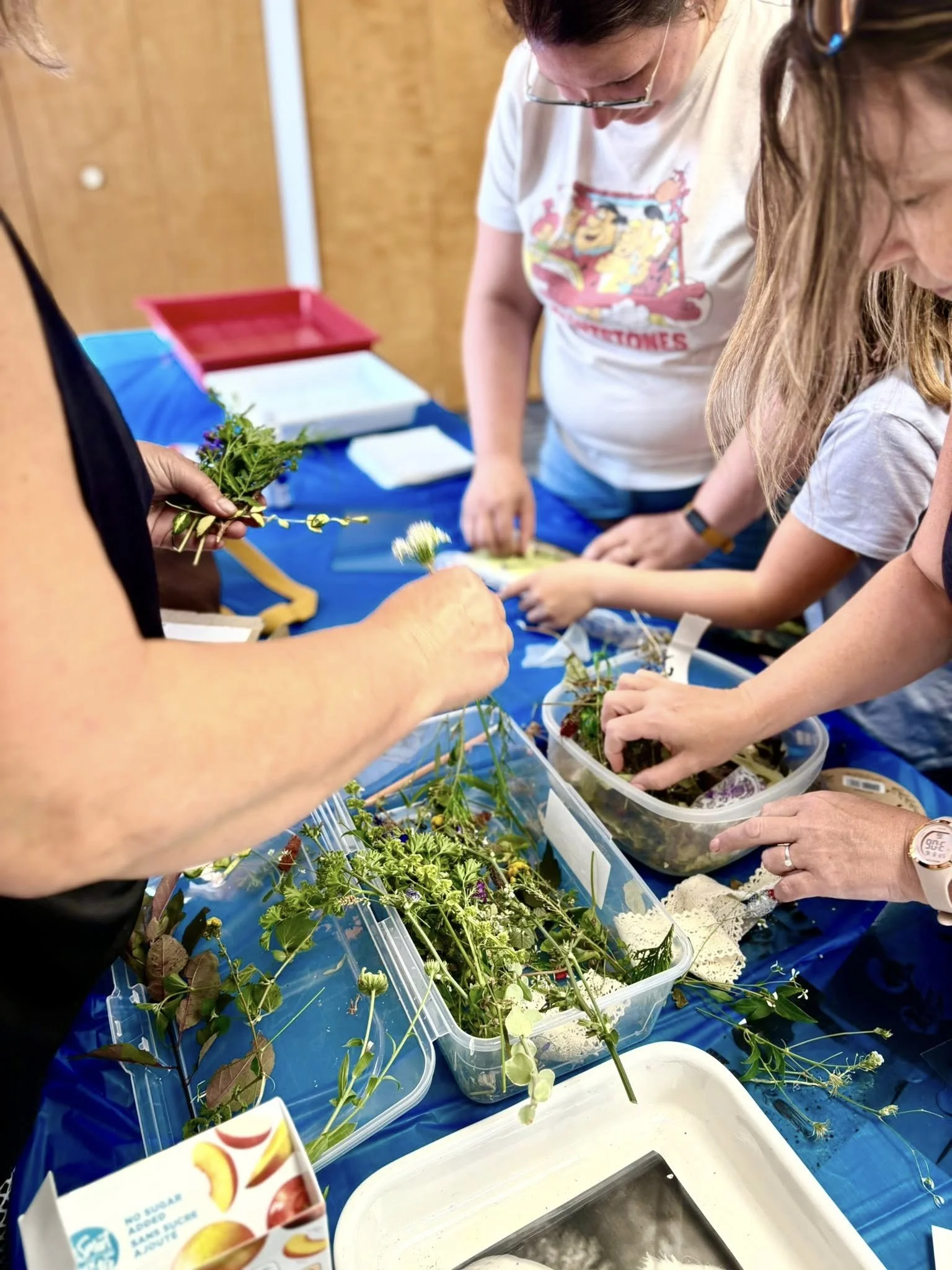 Groupe de personnes cueillant et manipulant des plantes ou des fleurs sur une table bleue, avec des fruits en carton en avant-plan.