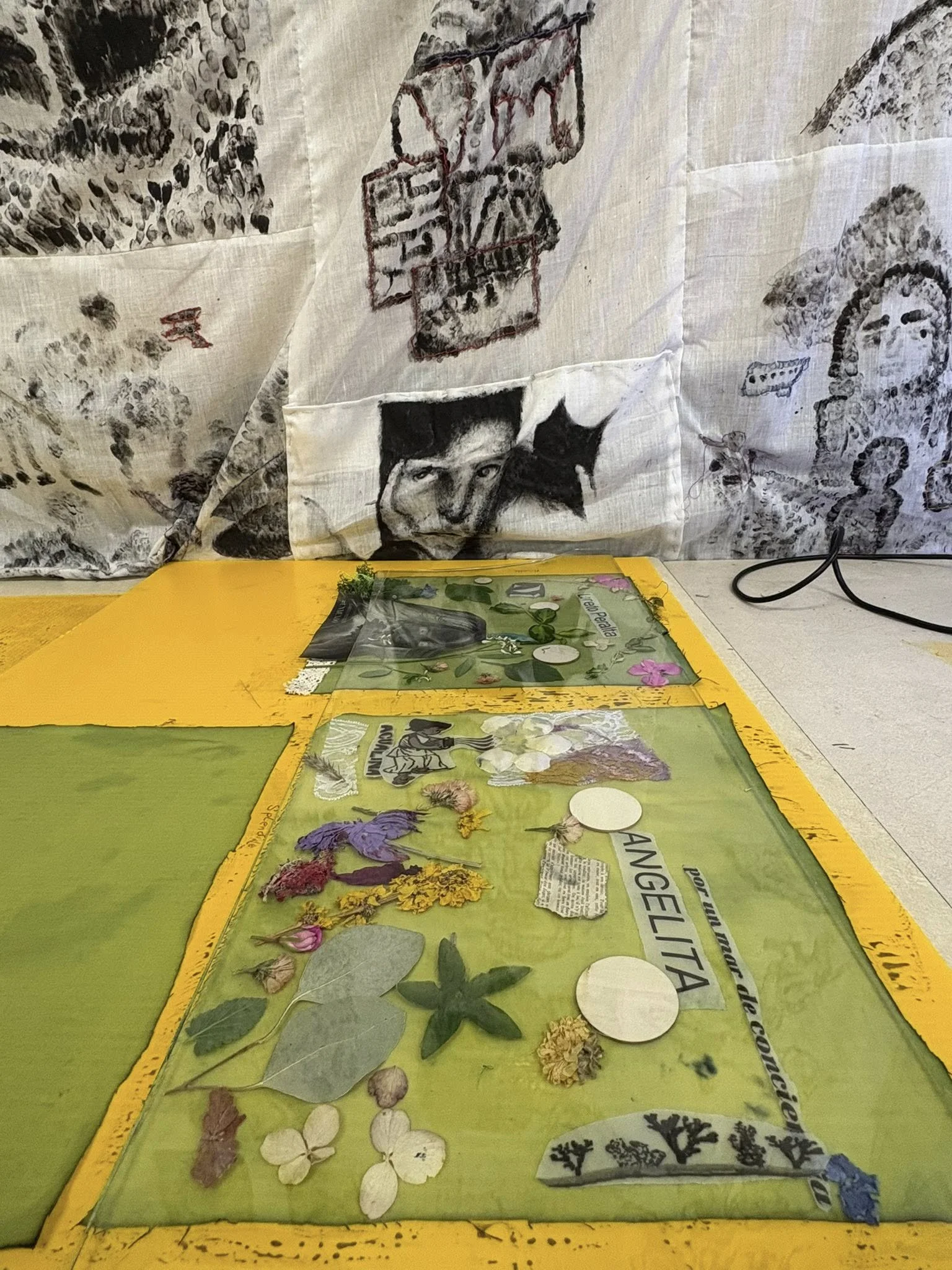 Une pièce avec une table jaune recouverte de plantes séchées, de photos et de papier, et un tissu en arrière-plan avec des dessins et inscriptions.
