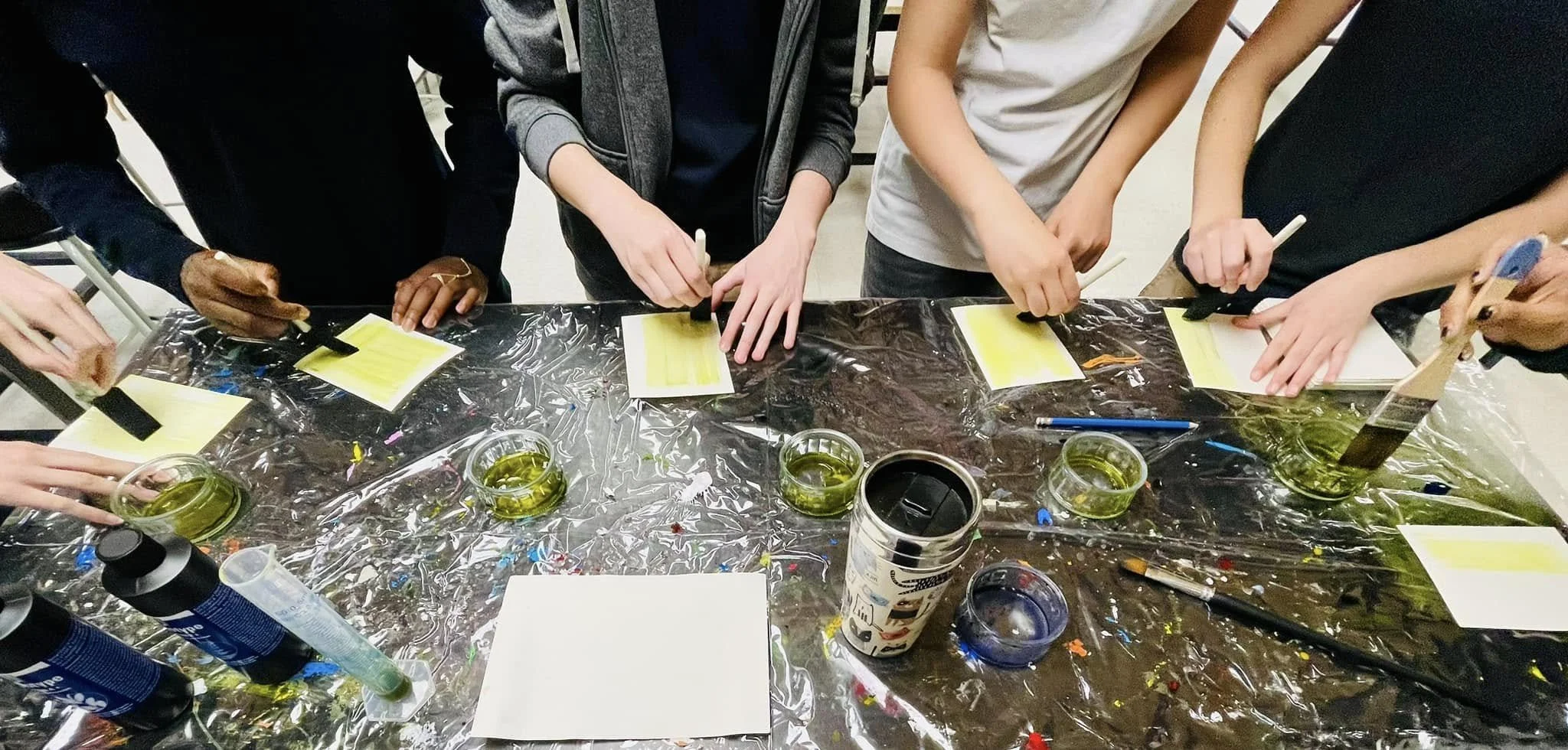 Groupe de personnes faisant de la peinture avec des pinceaux et des feuilles de papier, sur une table recouverte de plastique avec des pots de peinture verte.