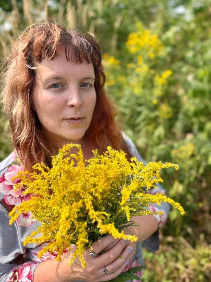 Une femme aux cheveux roux et aux yeux bleus tient un bouquet de fleurs jaunes dans un jardin. Elle porte un vêtement imprimé floral et a un piercing au nez et au labret.