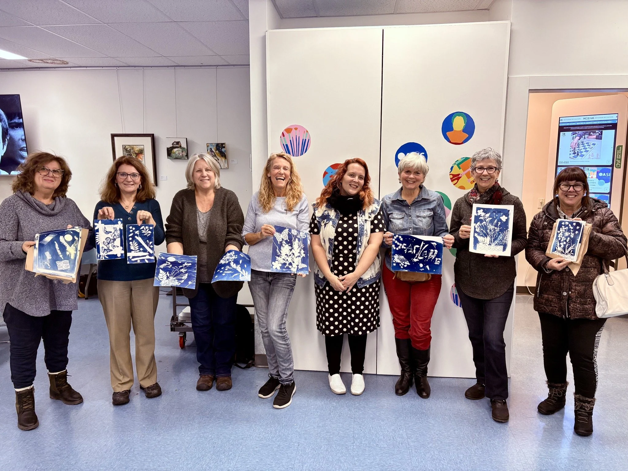 Groupe de neuf femmes posant dans une galerie d'art, chacune tenant ou montrant une pièce d'art en technique de cyanotype ou sérigraphie. Elles sourient, vêtues de vêtements décontractés, devant un mur blanc avec des œuvres d'art coloriées et un écra