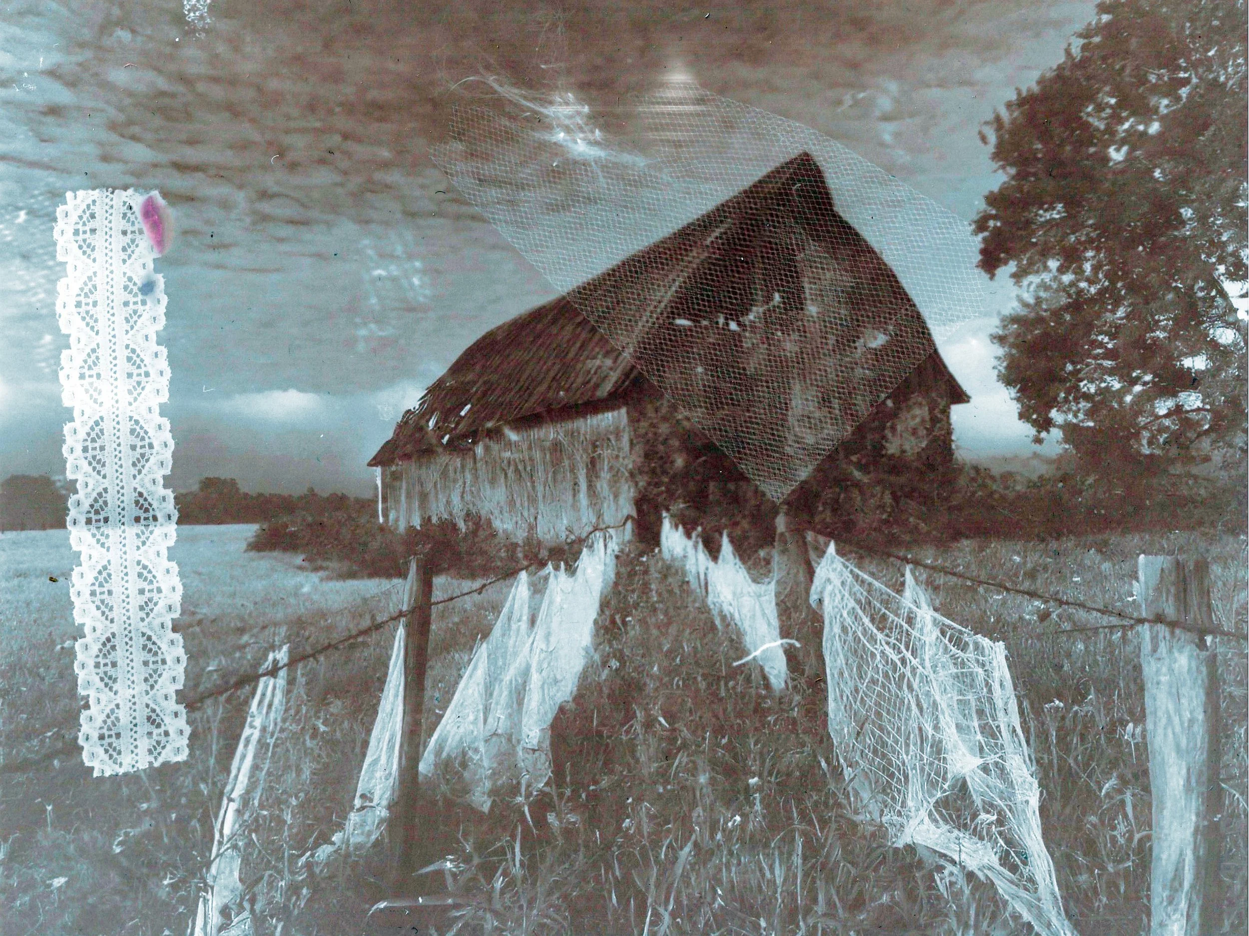 Ancienne ferme en bois avec un toit en chaume, entourée d'une clôture avec de vieux morceaux de tissus ou de dentelle suspendus dessus, dans un champ avec des arbres en arrière-plan, sous un ciel nuageux.