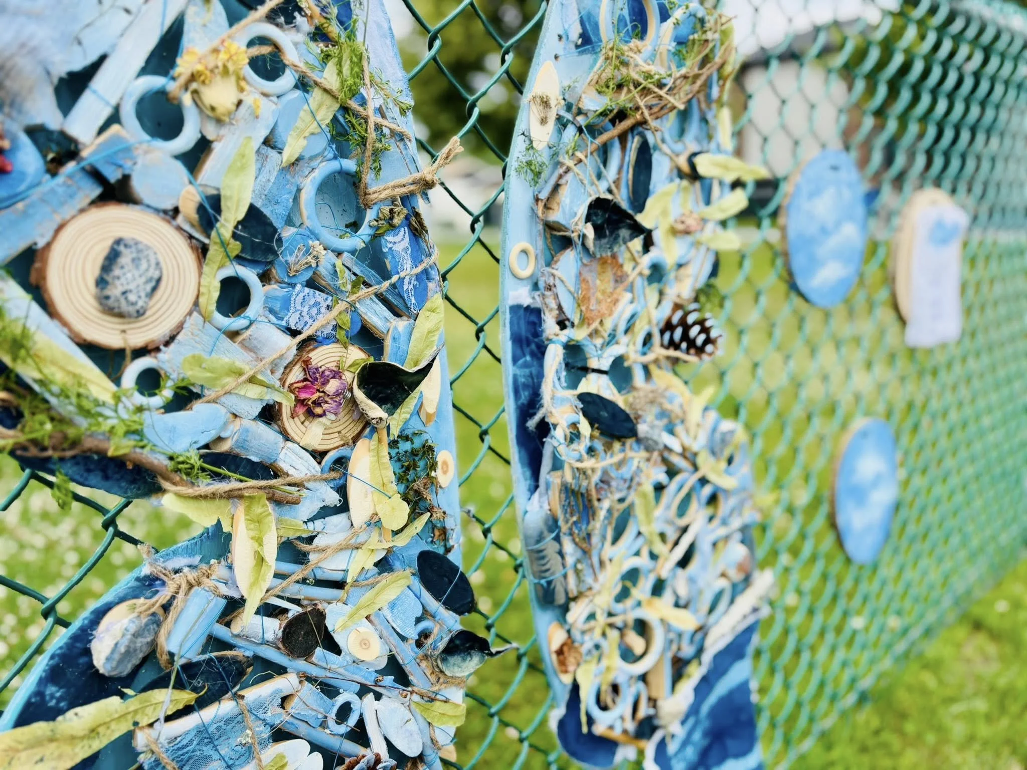 Décor de carré avec des morceaux de bois, de tissu, des coquillages, des feuilles, des champignons et d'autres objets attachés à une clôture en métal vert, avec un arrière-plan de gazon et de végétation.