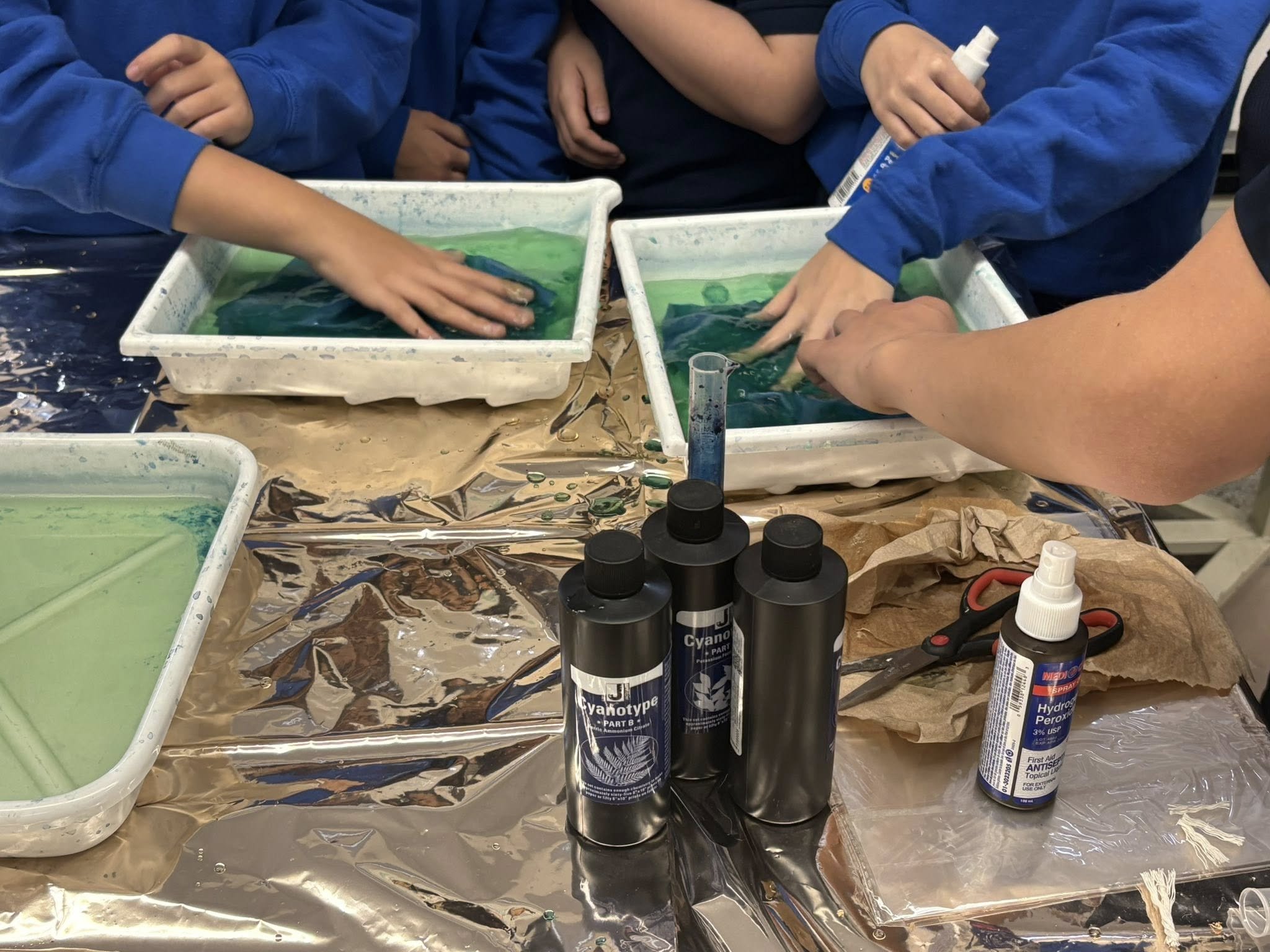Enfants en train de faire une expérience de mélange de liquides colorés dans des contenants en plastique, avec des bouteilles de produits chimiques et des ciseaux sur la table.