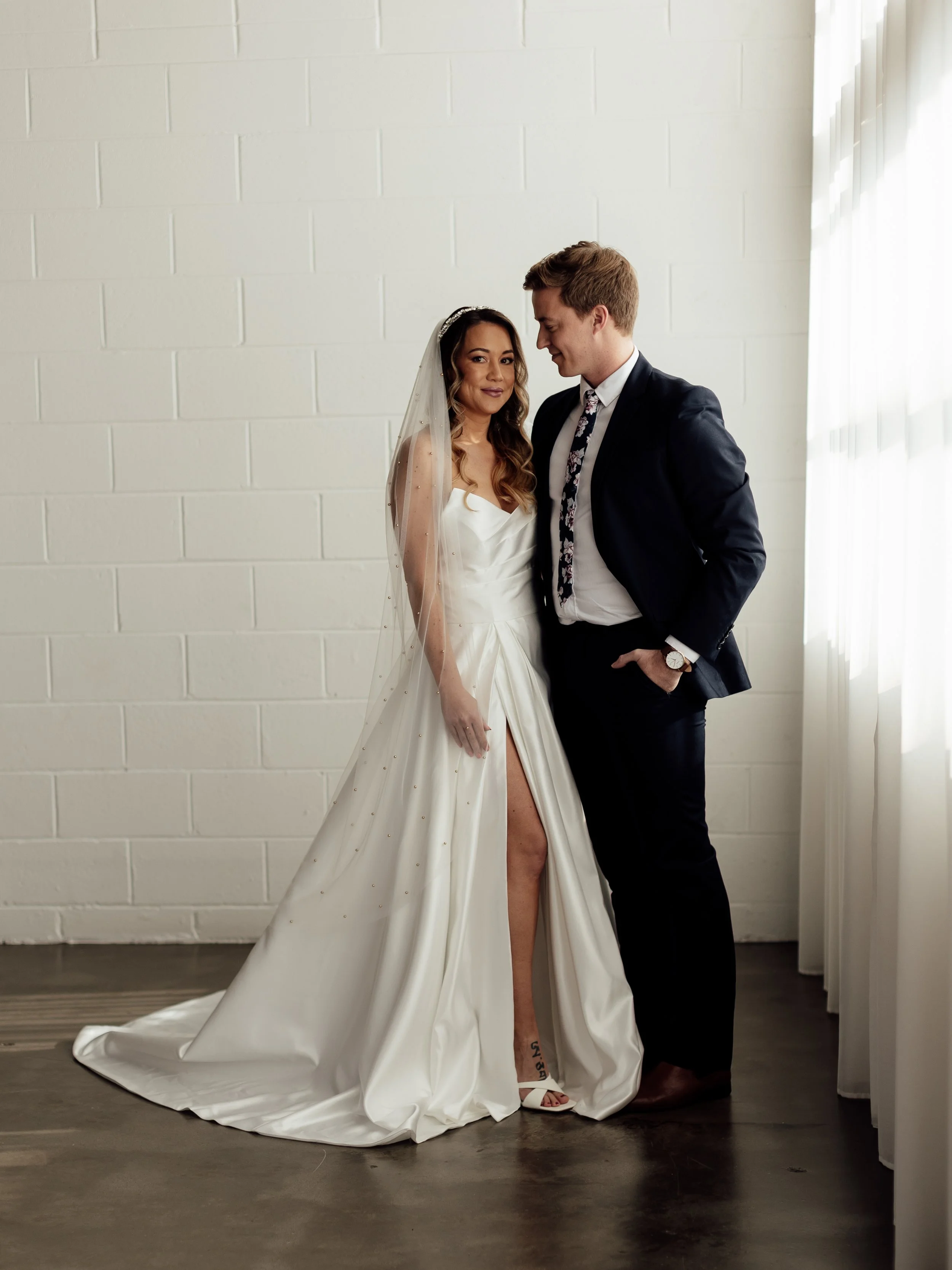 A bride and groom stand close together indoors, with the bride wearing a white wedding dress and veil, and the groom in a black suit, white shirt, and patterned tie. The bride has long wavy hair, and the groom has short light brown hair. They are smiling and looking at each other in front of a plain white brick wall with sunlight filtering through a window.