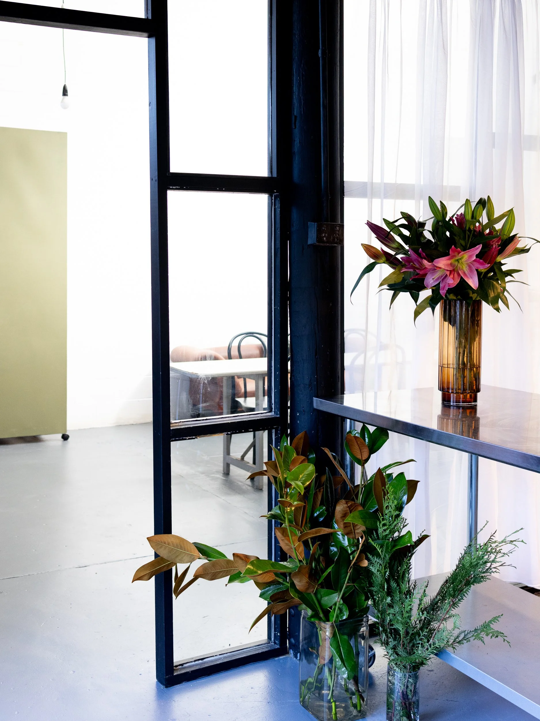 Interior view of a room with a black metal door frame, a window with white curtains, and various potted plants including lilies in a tall amber vase, a rubber plant, and other greenery.