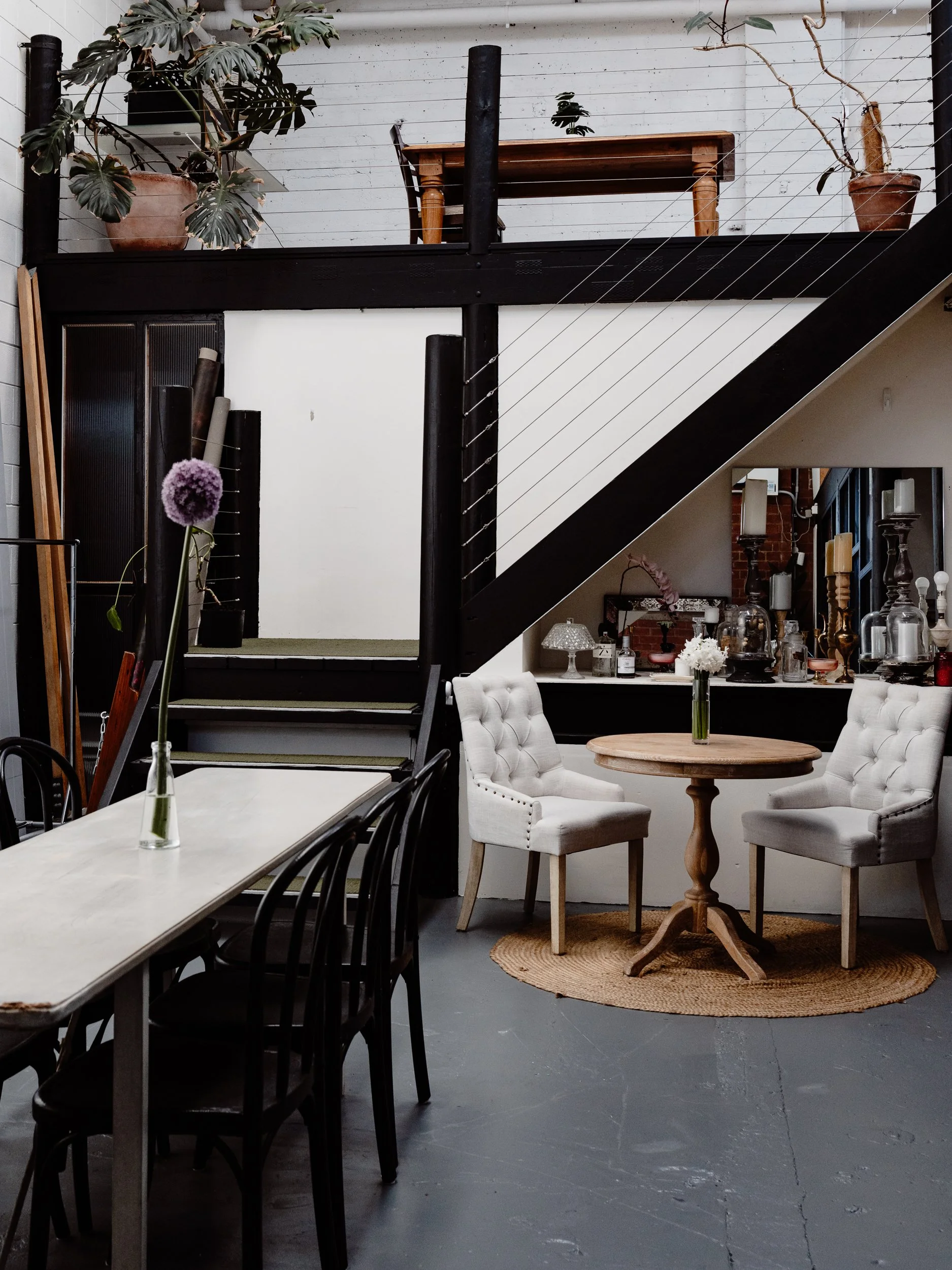 Interior of a room with a staircase, a round wooden table, and two white upholstered armchairs. The upper balcony has potted plants and a wooden table, and the room features decorative items including candles and glass jars.