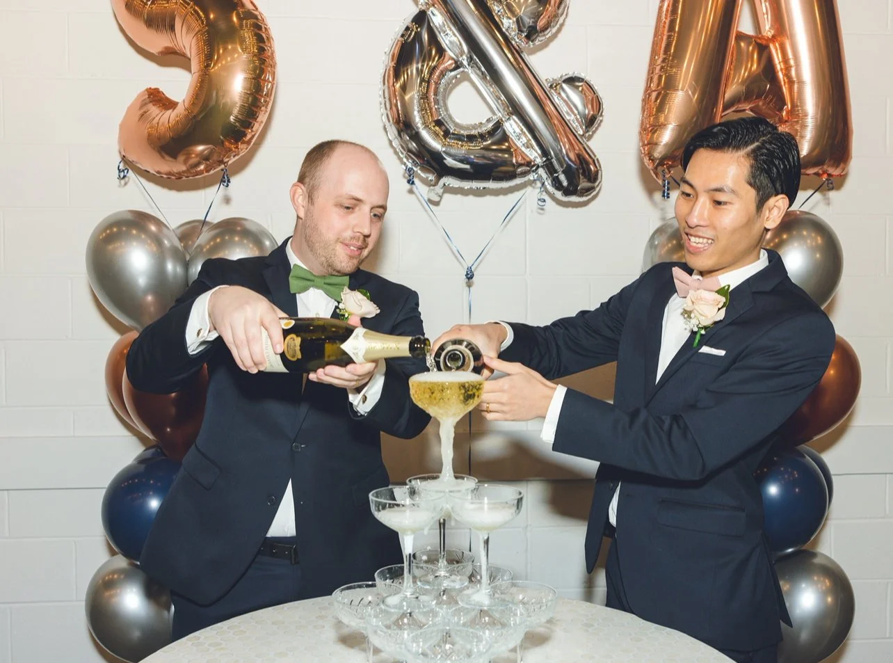 Two men in tuxedos with boutonnières pouring champagne into a tower of glasses at a celebration party with metallic balloons that spell out 2024 in the background.
