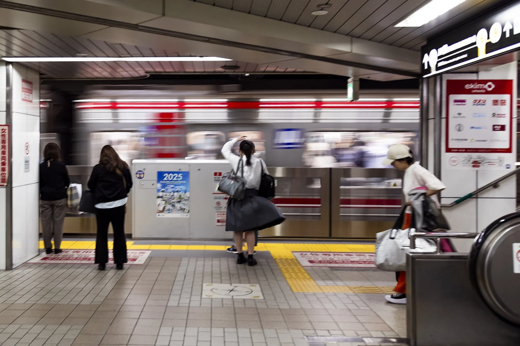 Scott Gould A Railway Life Photography an Osaka Metro train arriving at a station as people wait to join it. A girl holds her hair and her skirt flies as it is caught by the wind from the train.