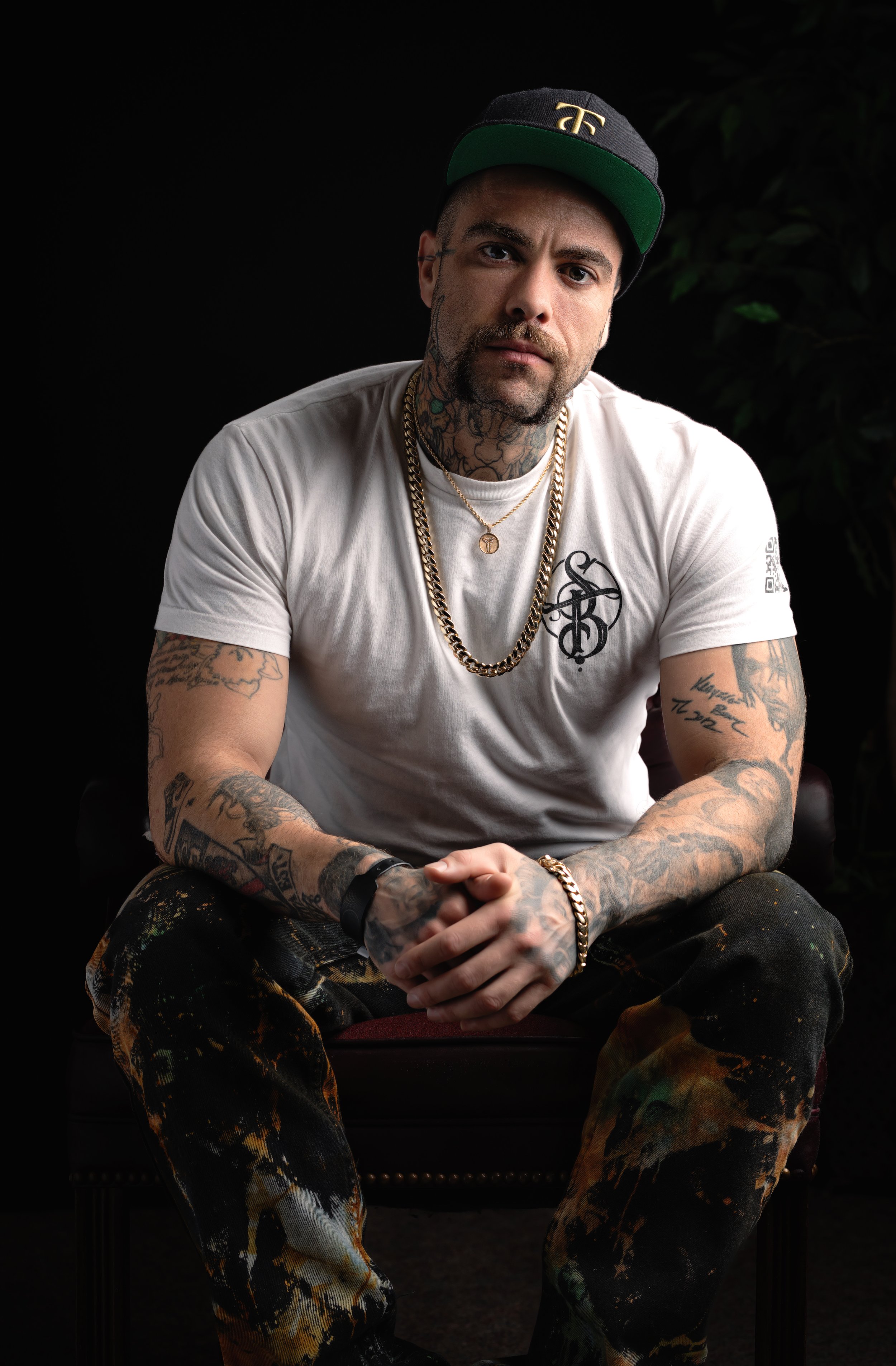 Man with tattoos wearing a white shirt and a black cap, sitting with hands clasped, against a dark background.