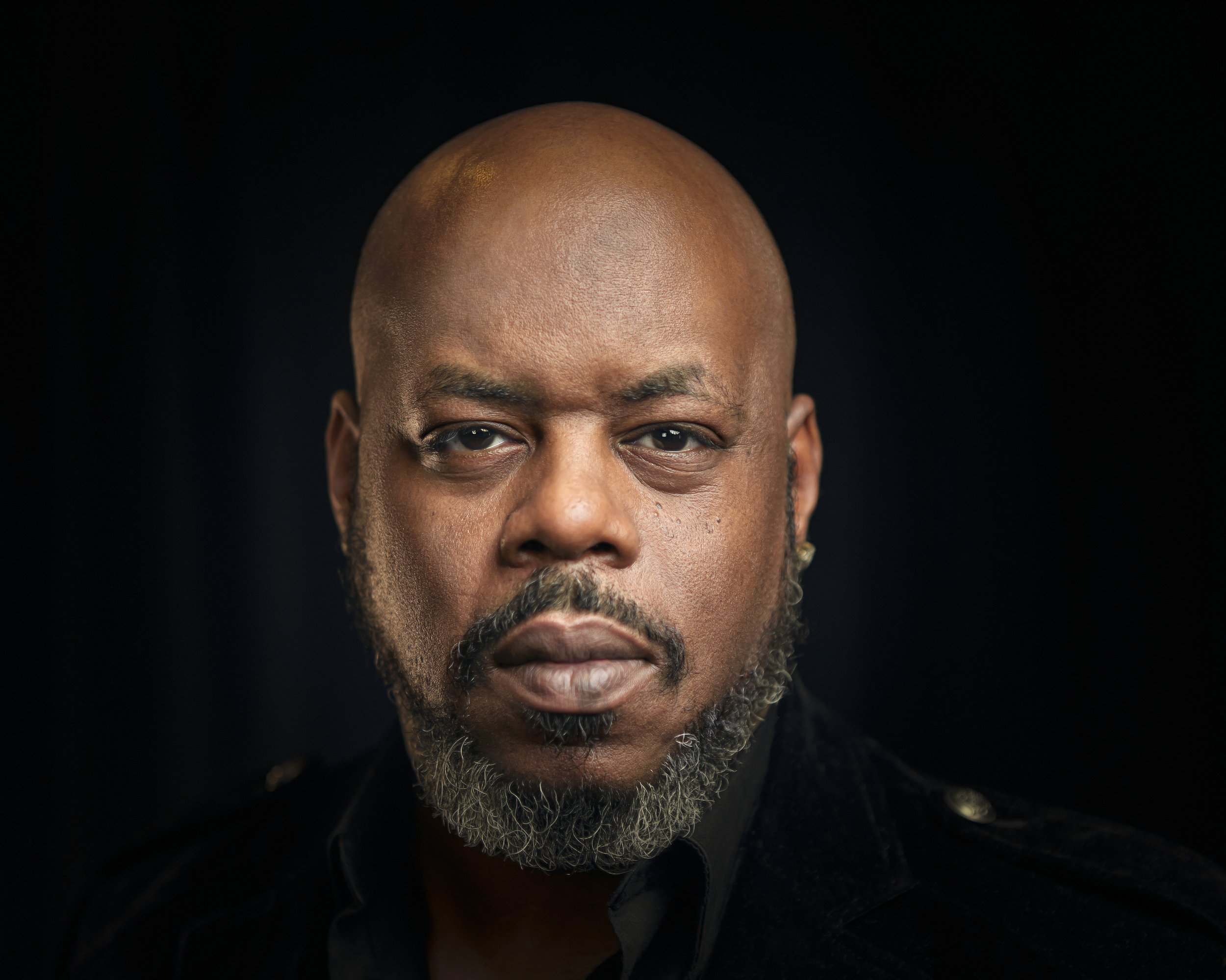 Portrait of a bald man with a beard looking directly at the camera against a dark background.